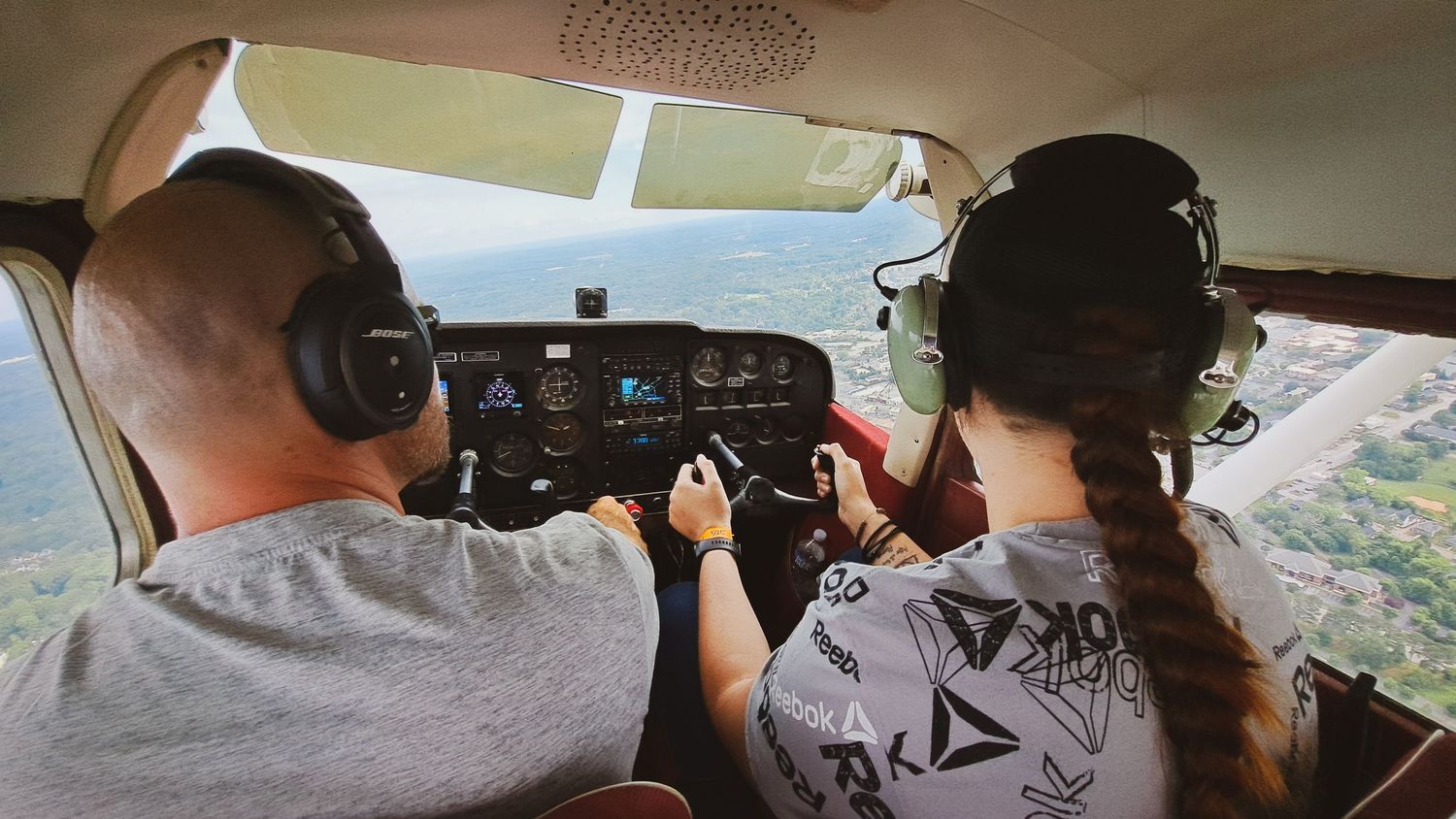 Two pilots wearing headsets fly a small aircraft over lush green landscape, viewed from behind inside the cockpit.