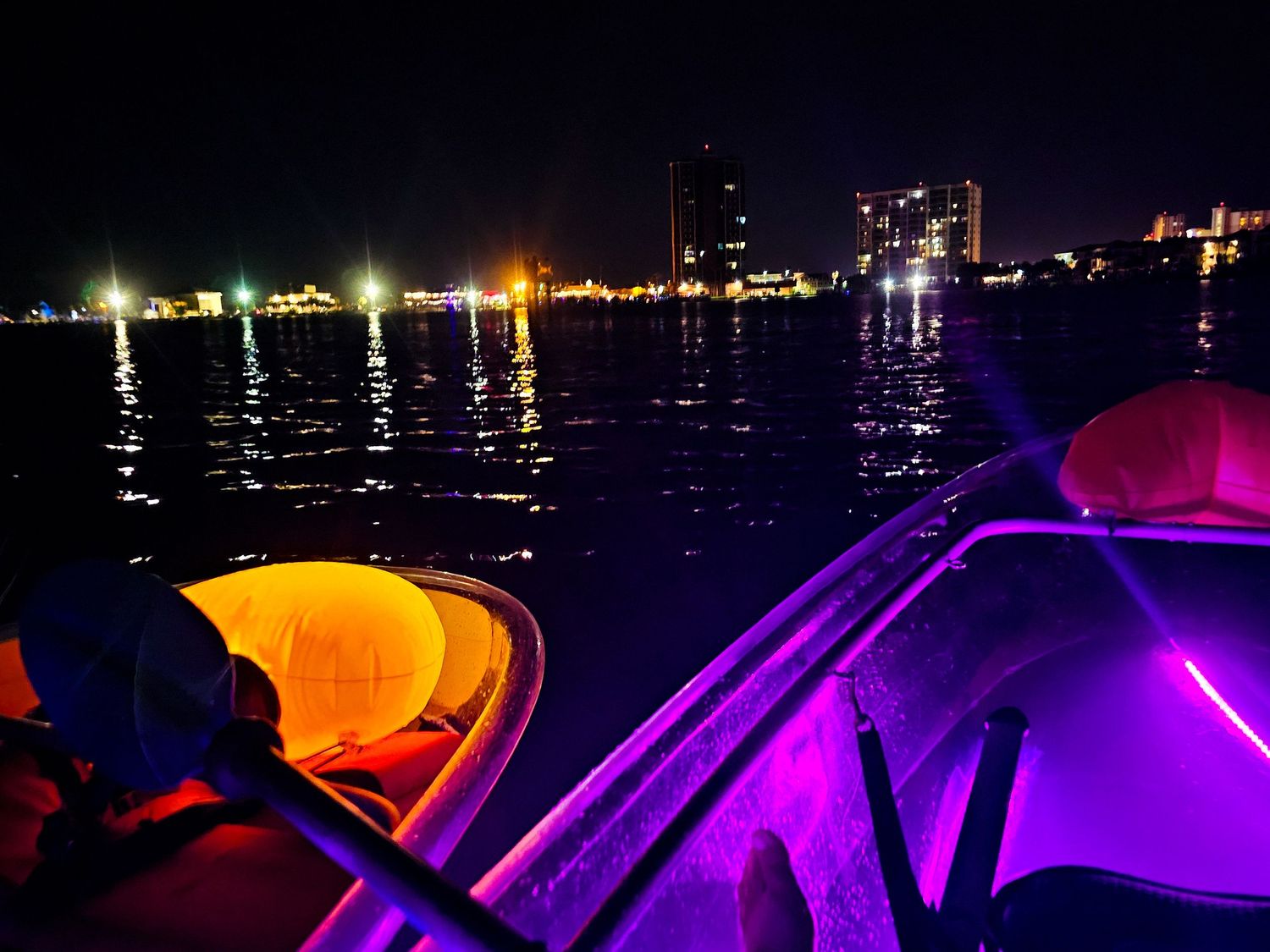 Illuminated boats glowing in purple and orange lights on dark water with city skyline at night.