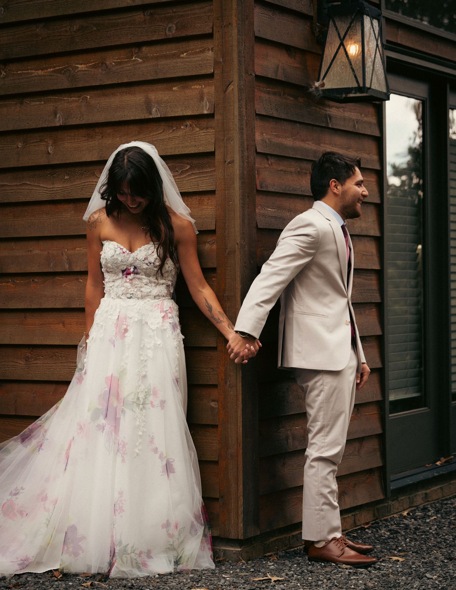 Bride and groom holding hands back-to-back outside rustic wooden building.