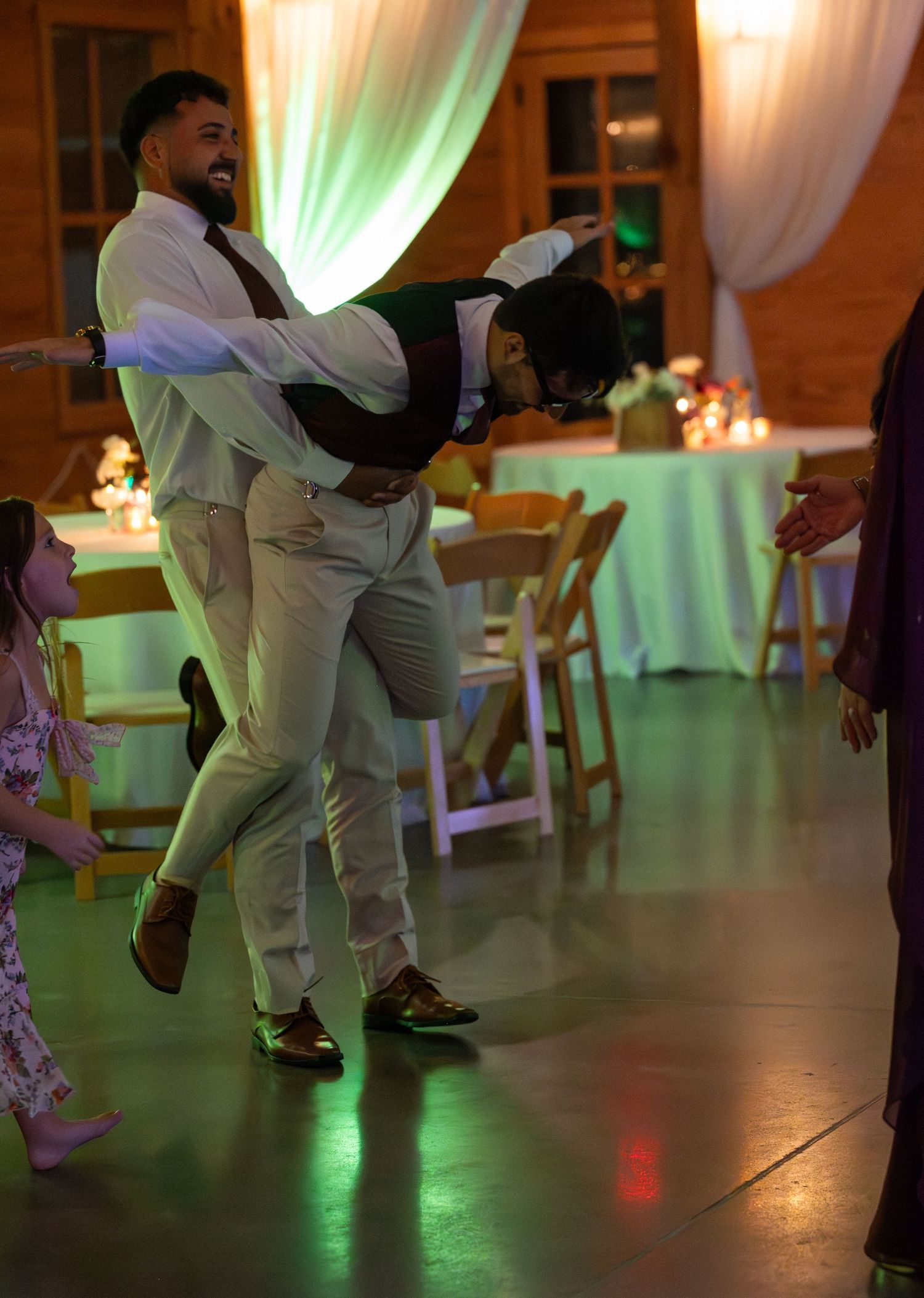 Two men joyfully dancing at a wedding reception in a decorated venue with green lighting and draped fabric.