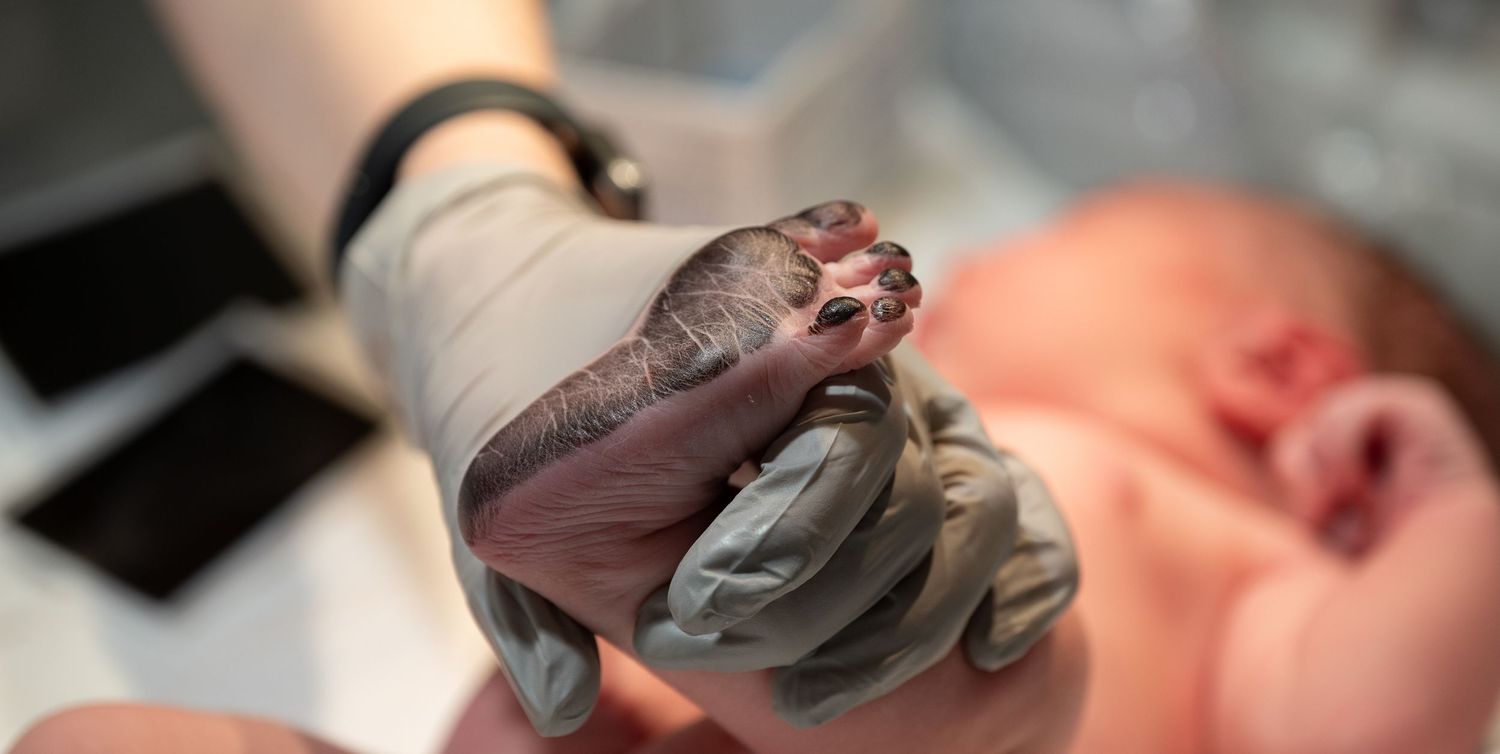 Gloved hands holding a newborn baby's tiny fist moments after birth in a medical setting.