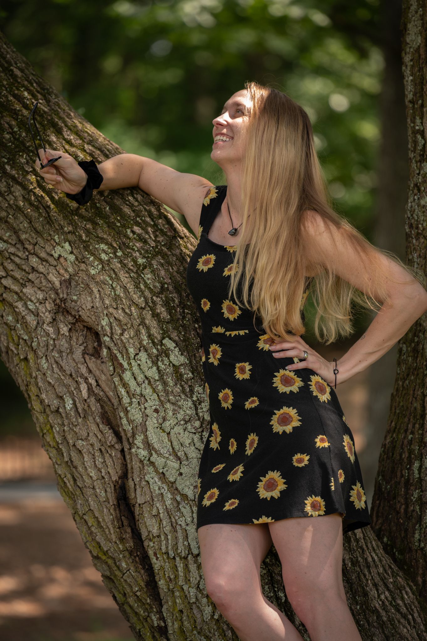 Smiling blonde woman in black sunflower dress poses against tree trunk in lush green forest setting.