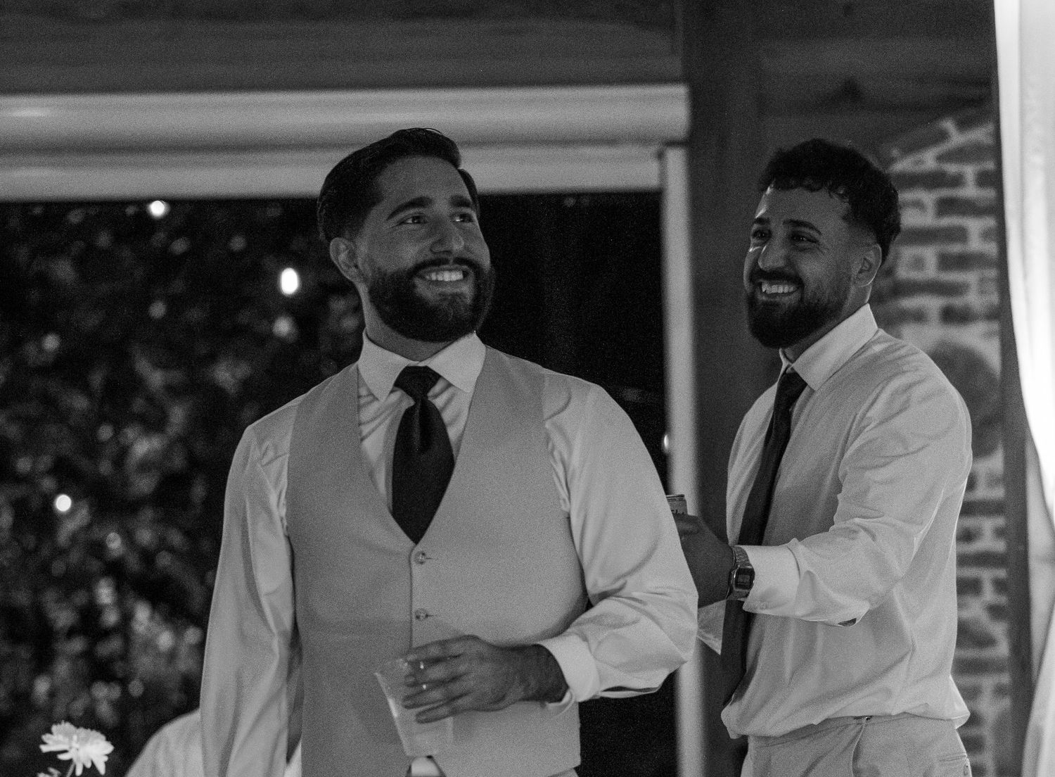 Two smiling groomsmen in white dress shirts and dark ties laugh together at a wedding reception.