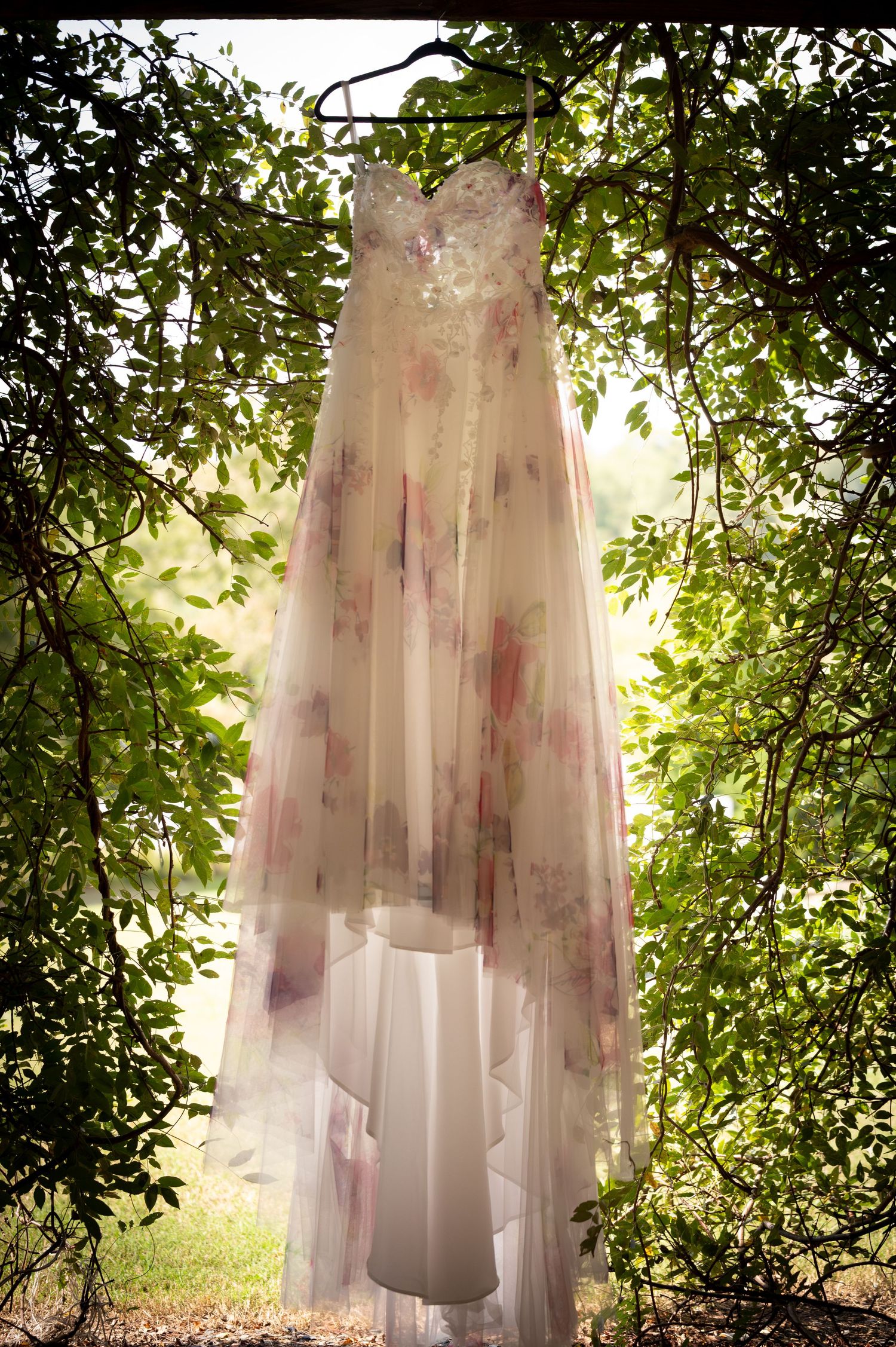 Floral wedding dress hanging on tree branches outdoors, featuring soft pink and purple watercolor blooms on sheer fabric.