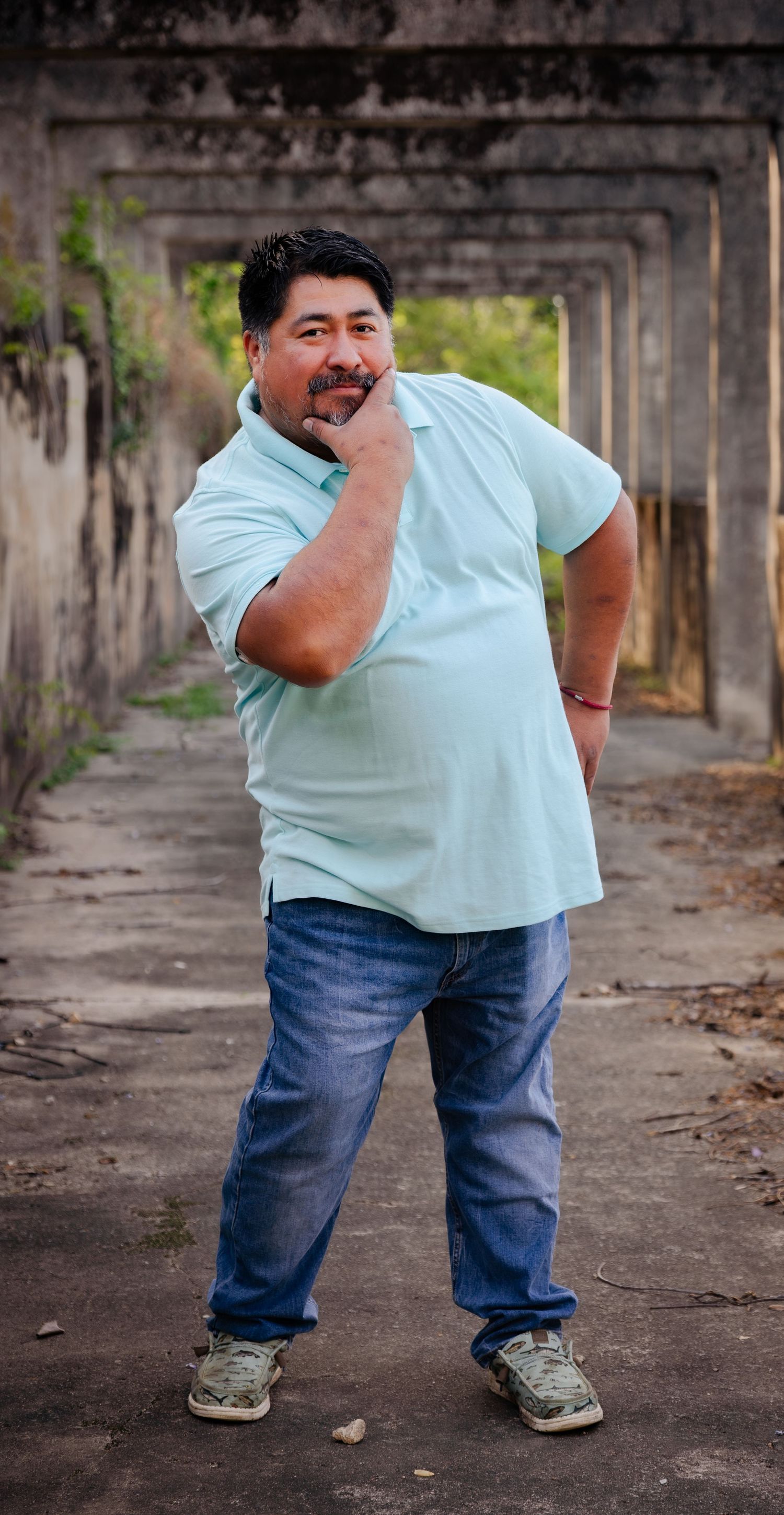 Heavyset Latino man in a light blue polo shirt and jeans posing confidently in an outdoor stone corridor.