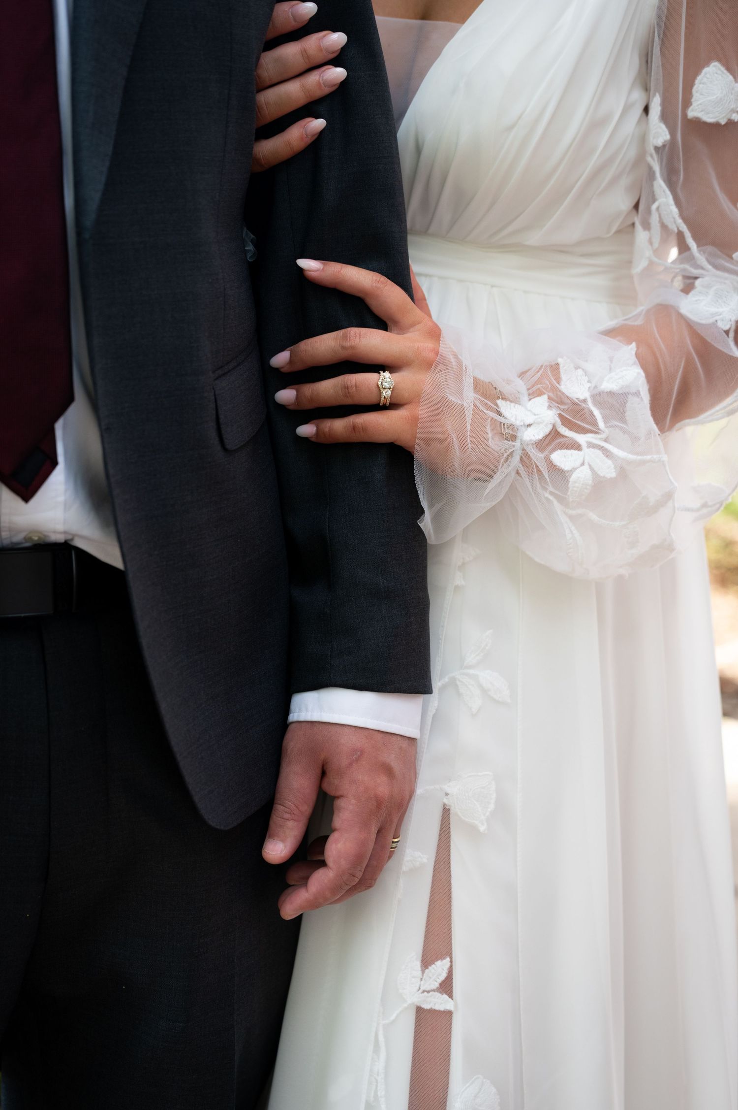 Close-up of bride and groom holding hands, showcasing wedding ring, white lace dress, and dark suit.