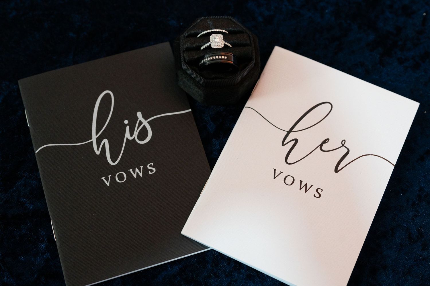 His and her wedding vow books with a diamond engagement ring resting on top, displayed on a dark velvet surface.