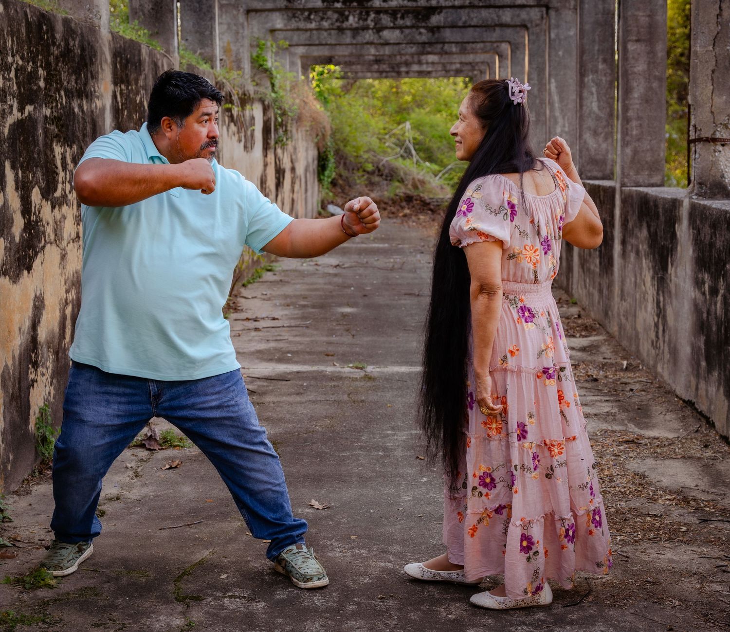 A man and woman playfully sparring in a rustic outdoor corridor, she in a floral dress, he in a blue shirt.