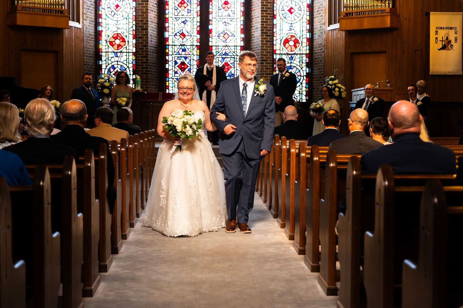 Bride and groom walk down church aisle after wedding ceremony, with stained glass windows and wooden pews in background.