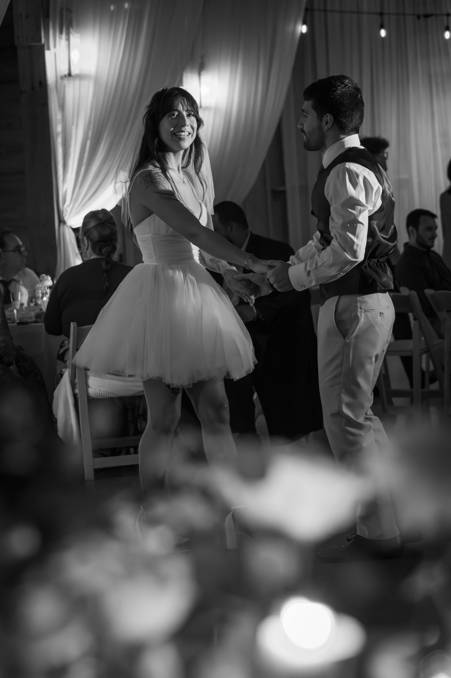 Couple sharing first dance at wedding reception in black and white photo, bride in short tulle dress, groom in vest.