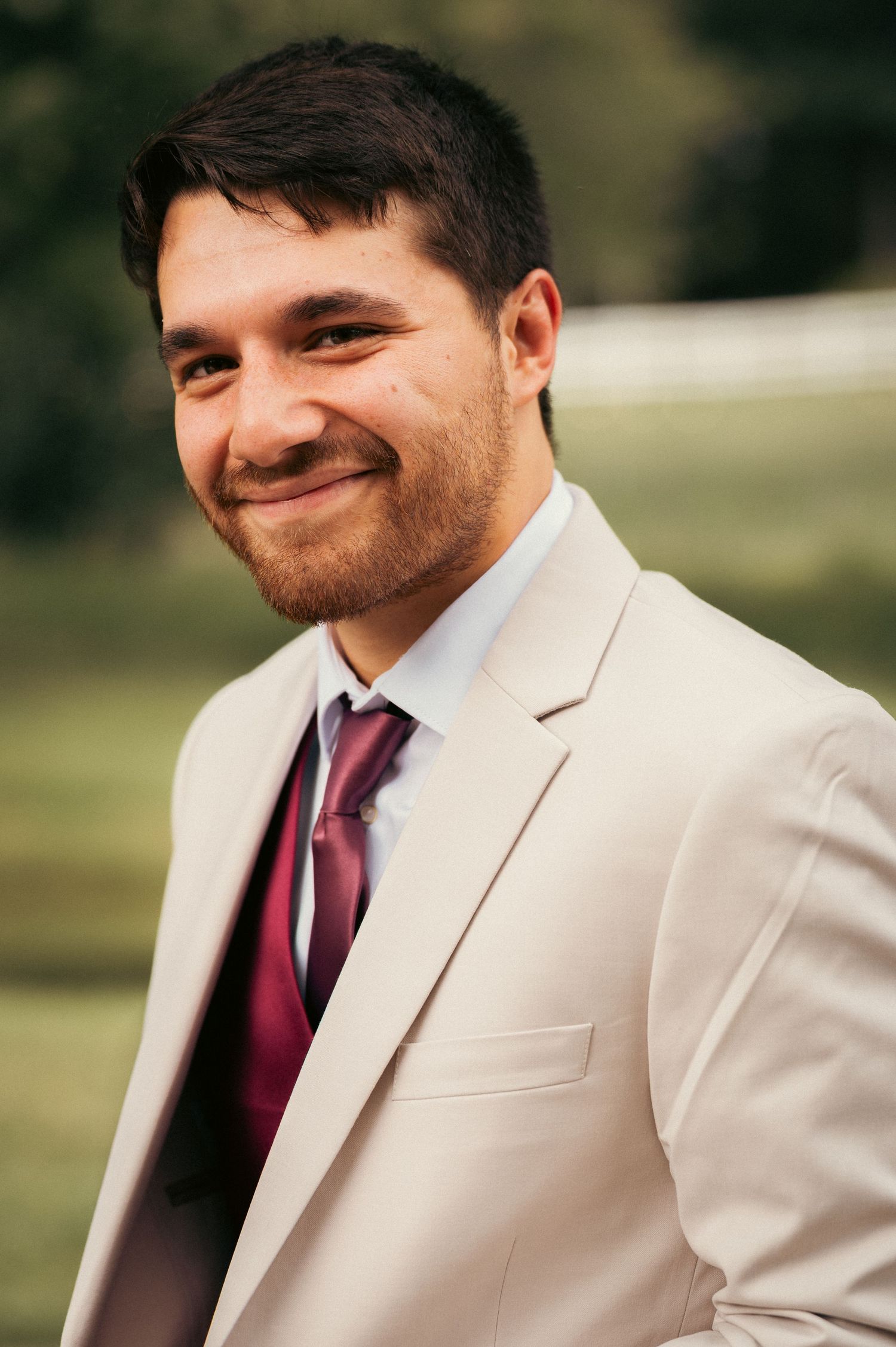 Smiling young man with beard wearing a cream suit and burgundy tie outdoors at a formal event.