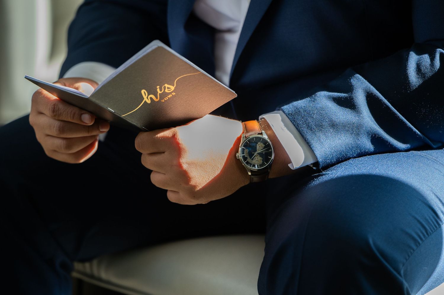 Man in navy suit holding a gold-scripted card and wearing a luxury watch, elegant wedding day detail.