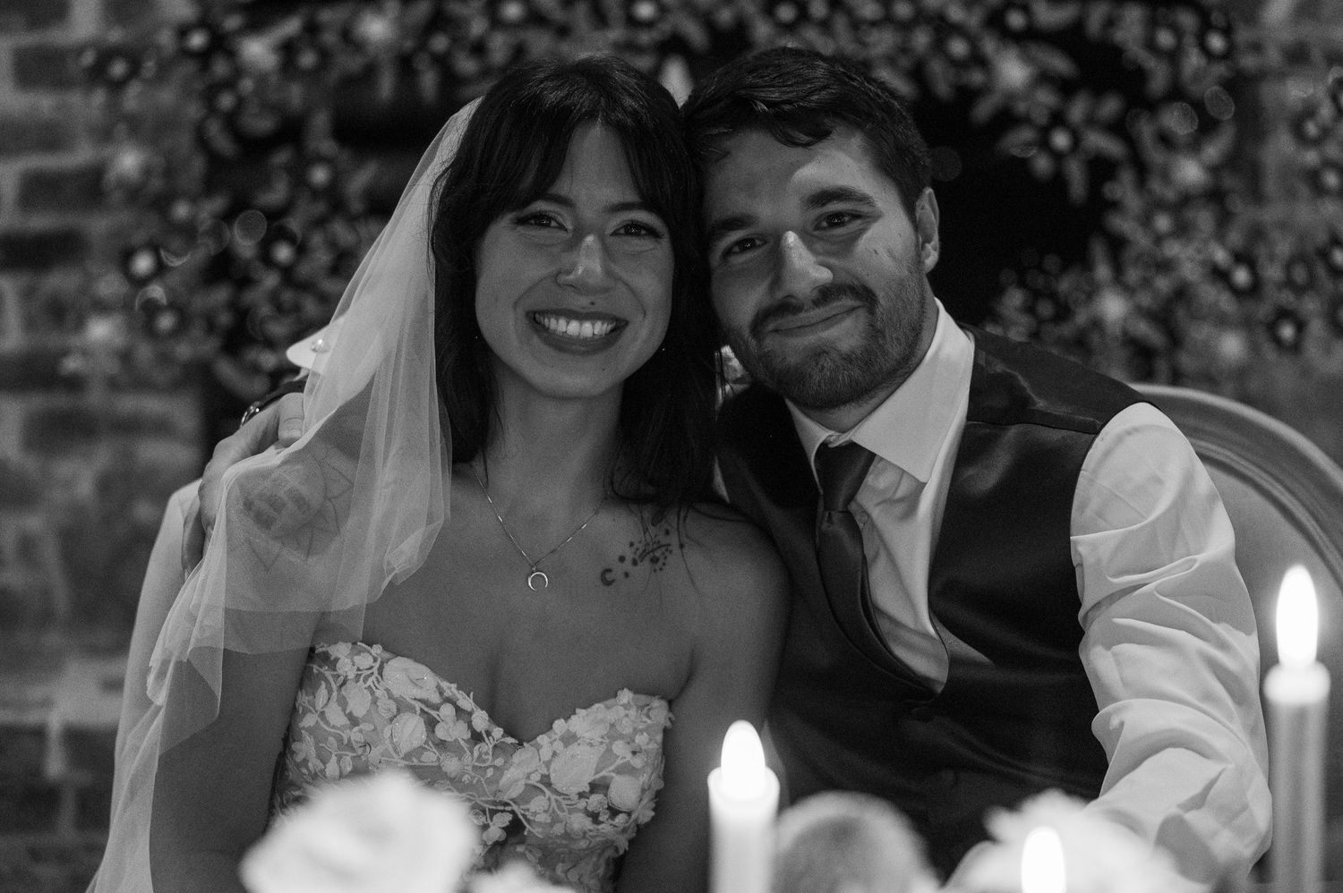 Black and white wedding photo of smiling bride in veil and groom in suit sitting together by candlelight.