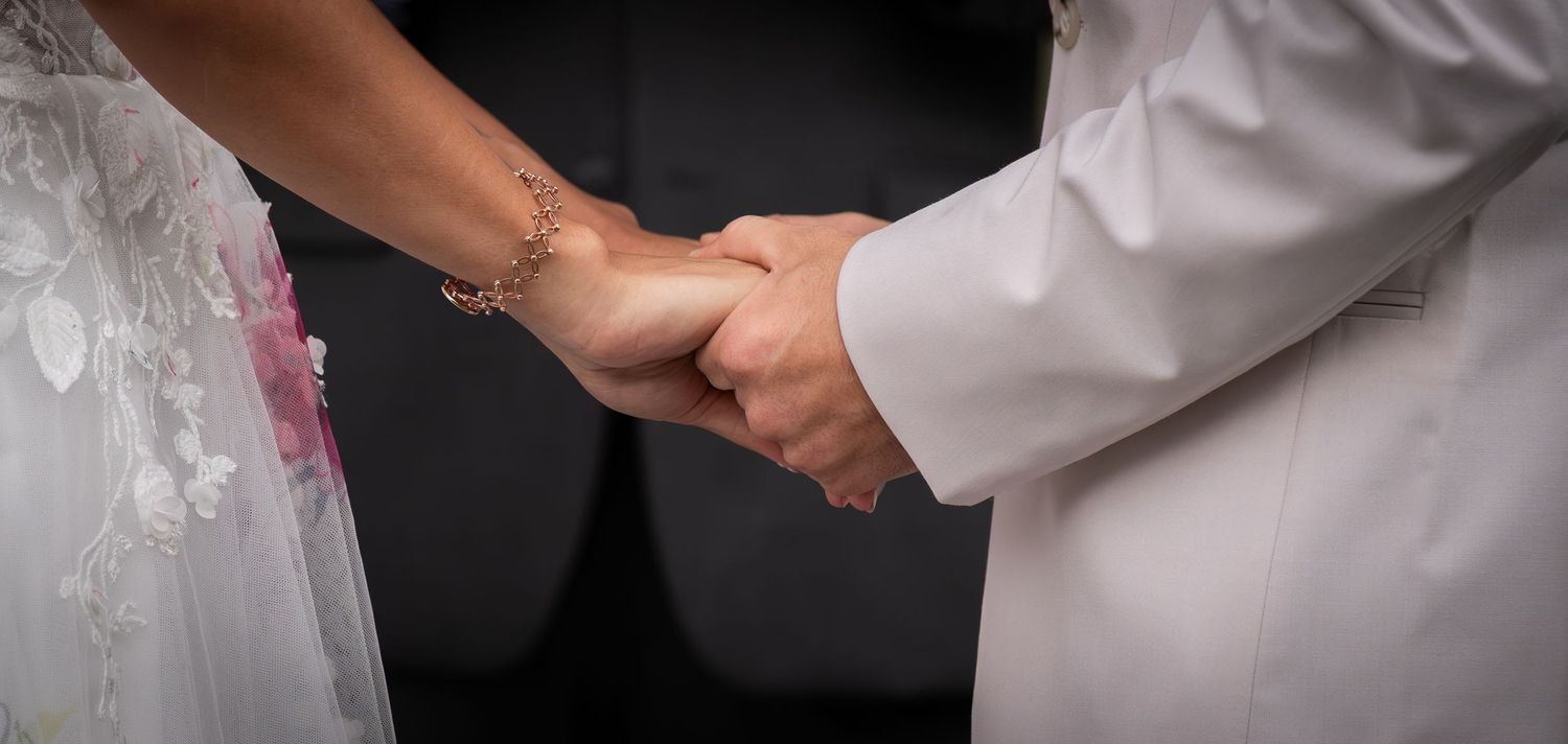 Close-up of bride and groom holding hands during wedding ceremony, bride wearing lace gown with bracelet.