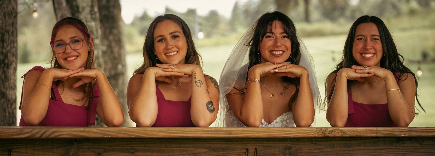 Four smiling women in pink bridesmaid dresses and bride posing together at wooden railing.