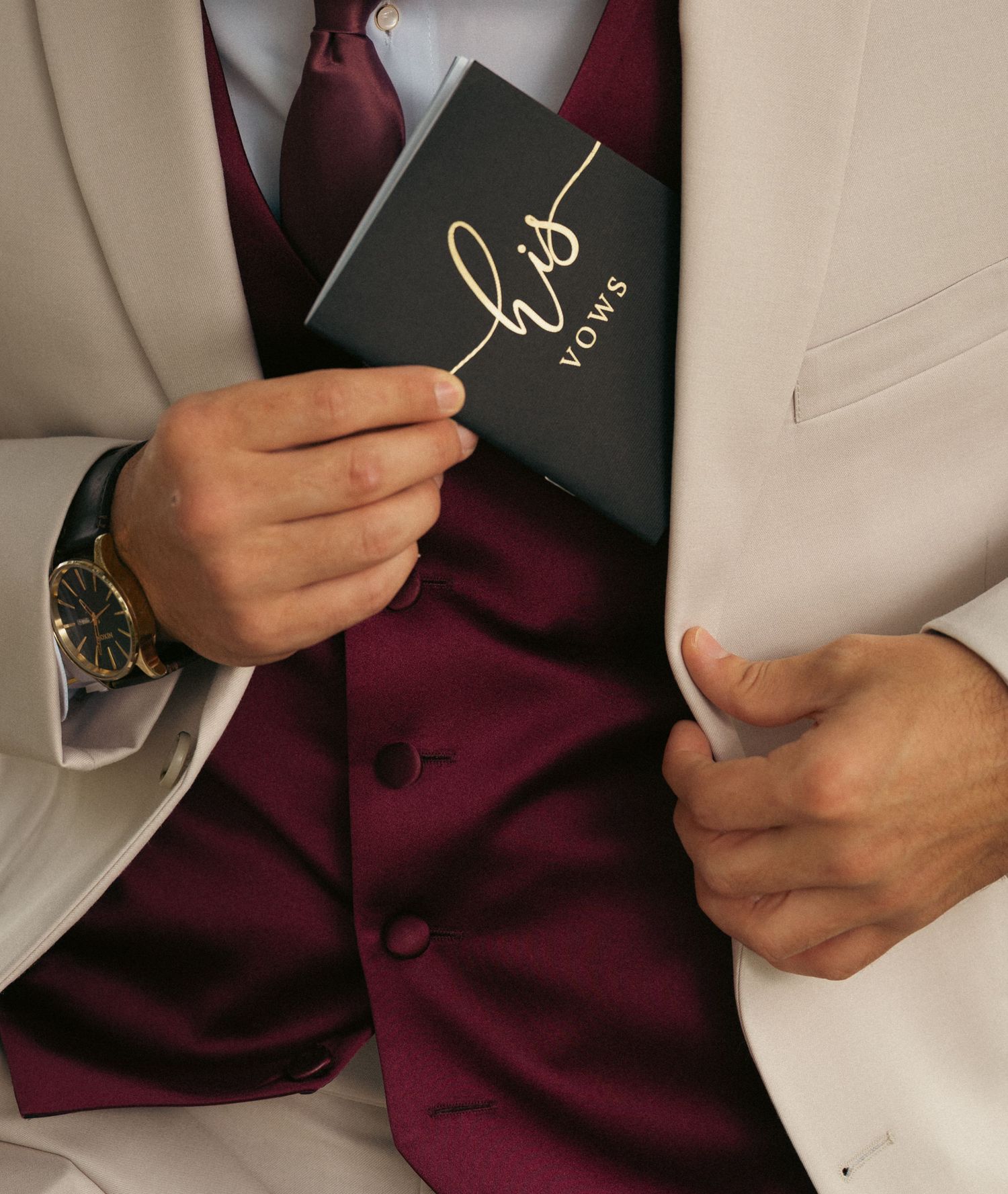Groom in white tuxedo with burgundy vest holding a black and gold 'His Vows' booklet on wedding day.