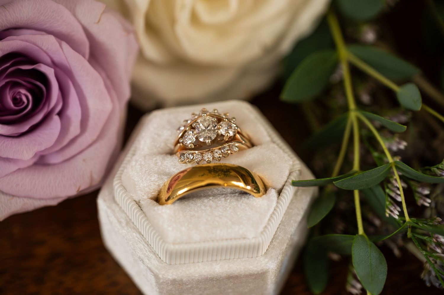 Gold wedding band and diamond engagement ring set displayed in a velvet box with roses and greenery.