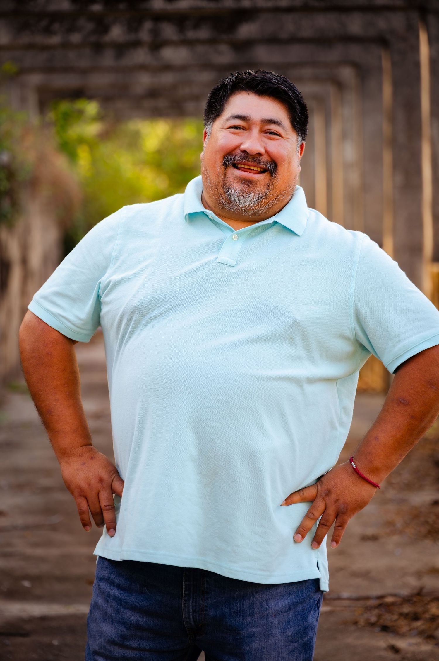 Smiling heavyset man in mint polo shirt standing confidently with hands on hips outdoors.
