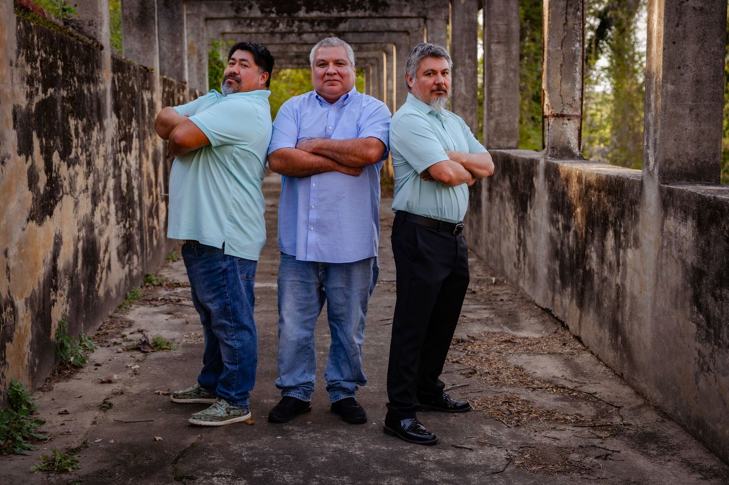 Three men stand with arms crossed in a rustic stone corridor, posing confidently for a portrait photo outdoors.