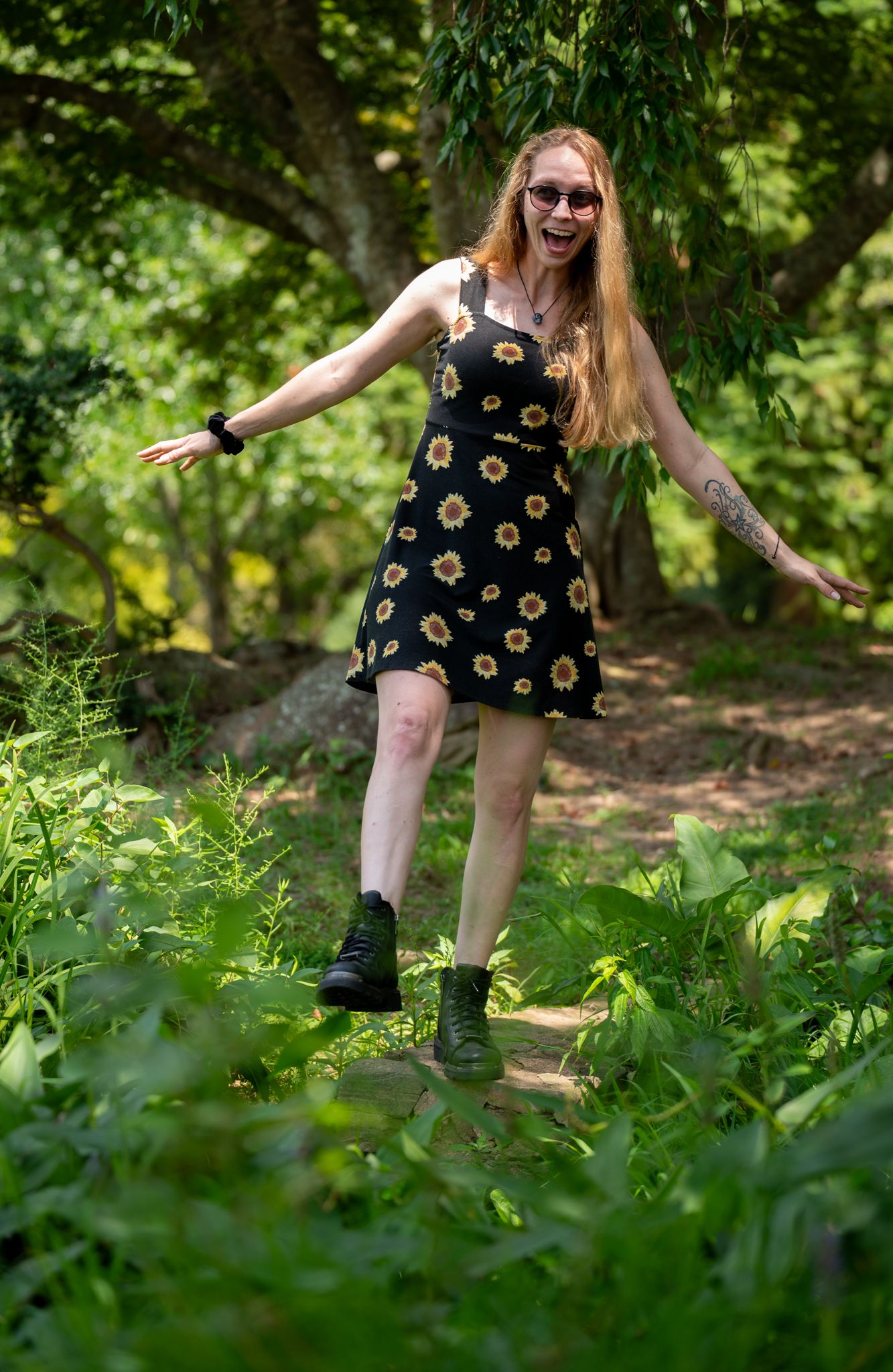 A smiling redhead woman in a black sunflower dress and combat boots balancing on a path in a lush green forest.