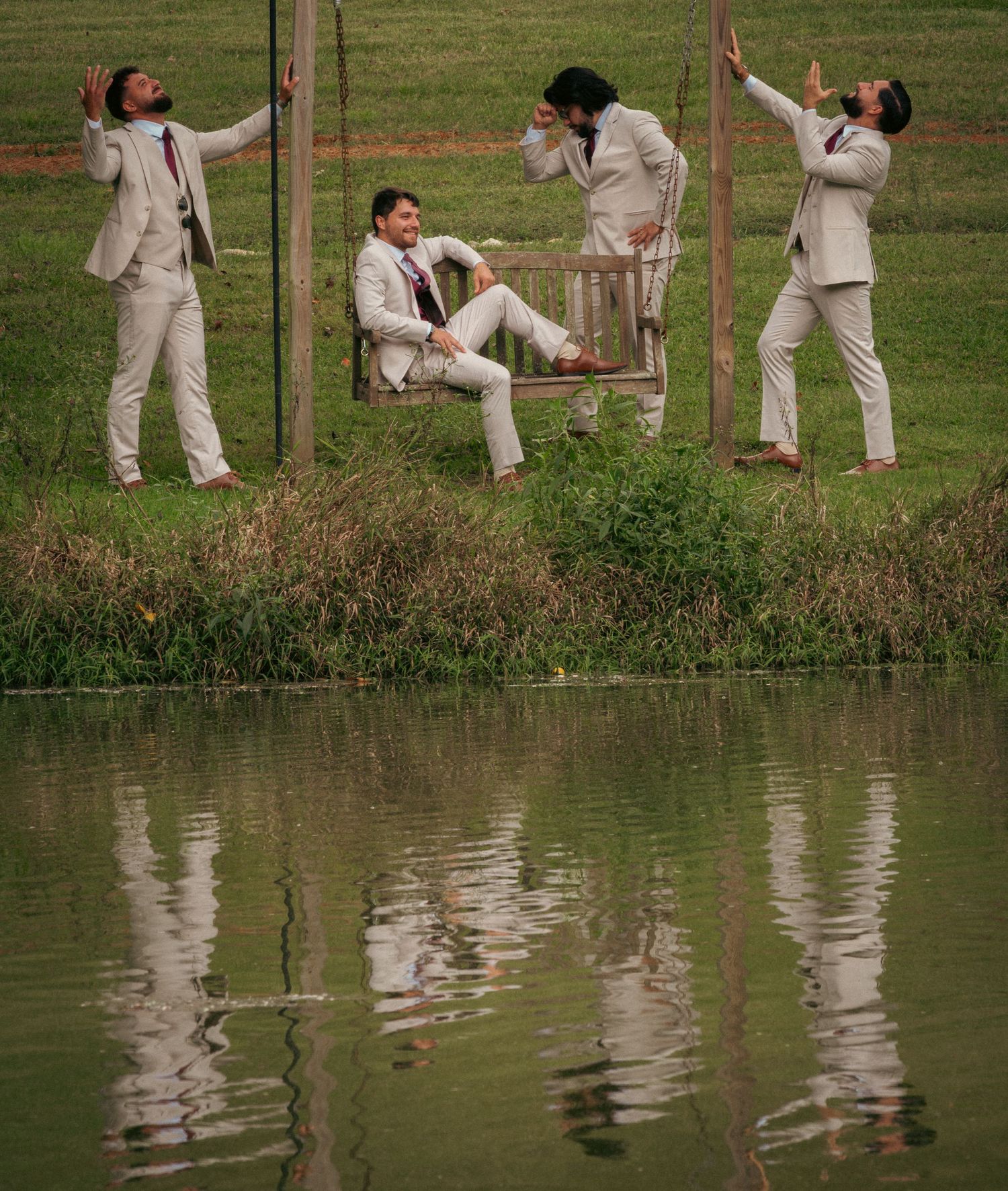 Four groomsmen in beige suits pose playfully near a pond with reflections in the water.