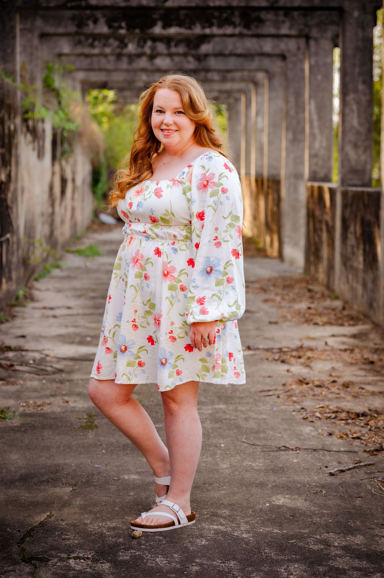 Young woman with red hair in a floral dress smiling outdoors in a rustic archway setting.