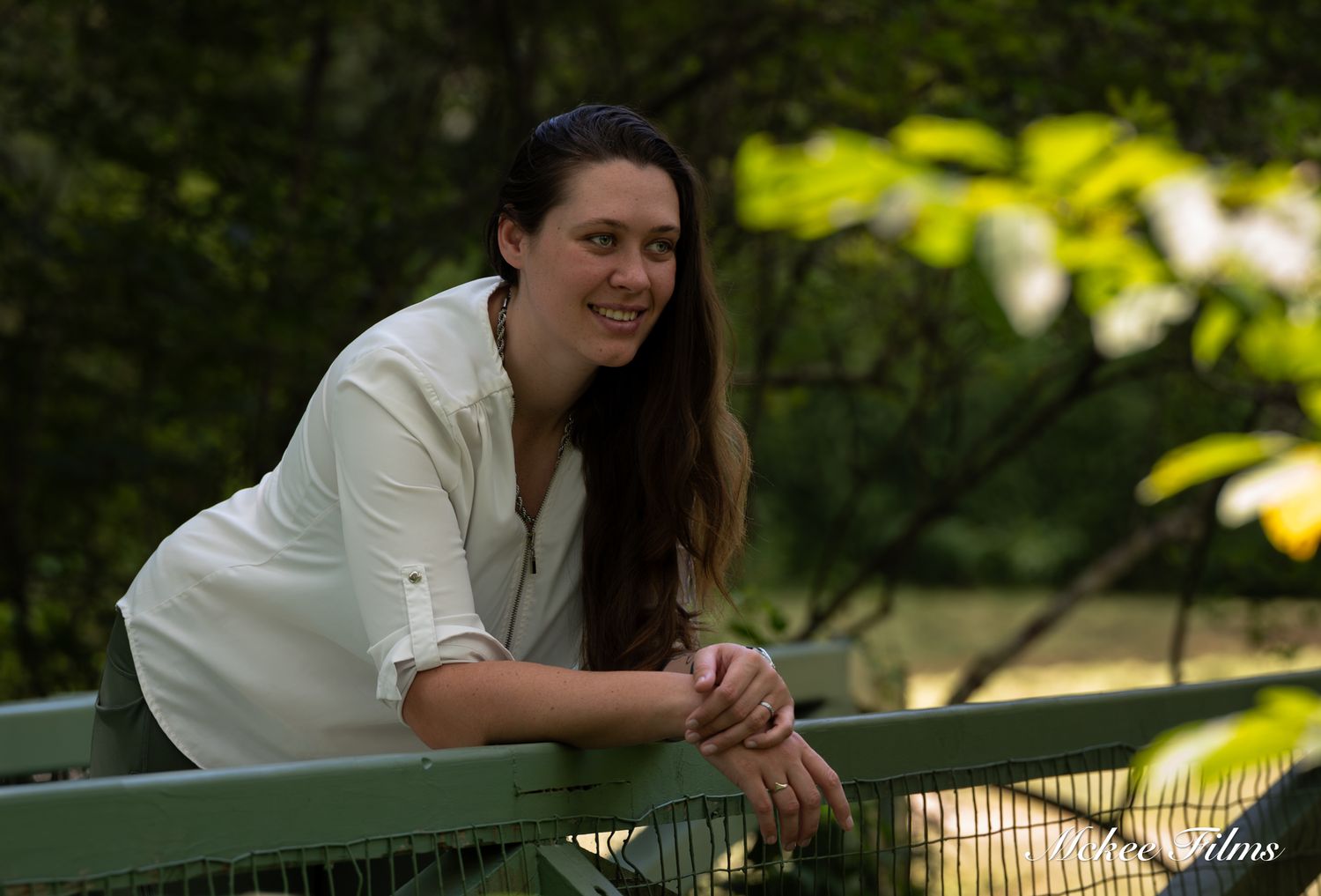 Smiling woman in white shirt leaning on green bridge railing outdoors.