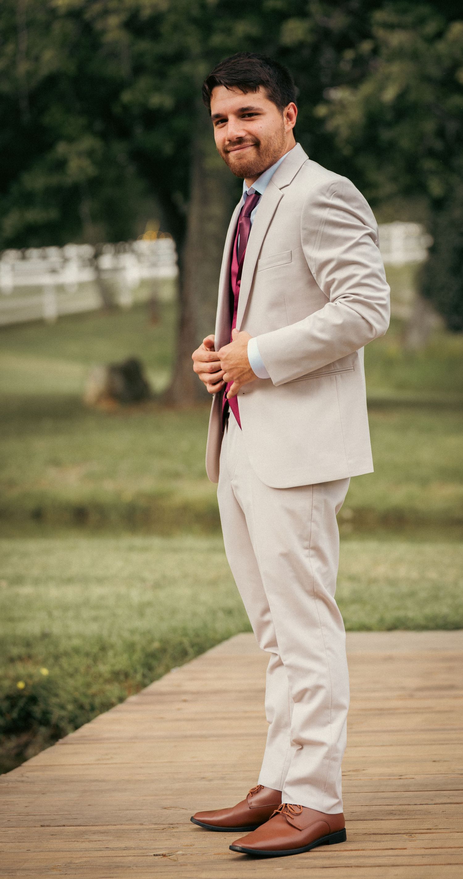 Smiling groom in beige suit with burgundy tie standing on wooden path outdoors.