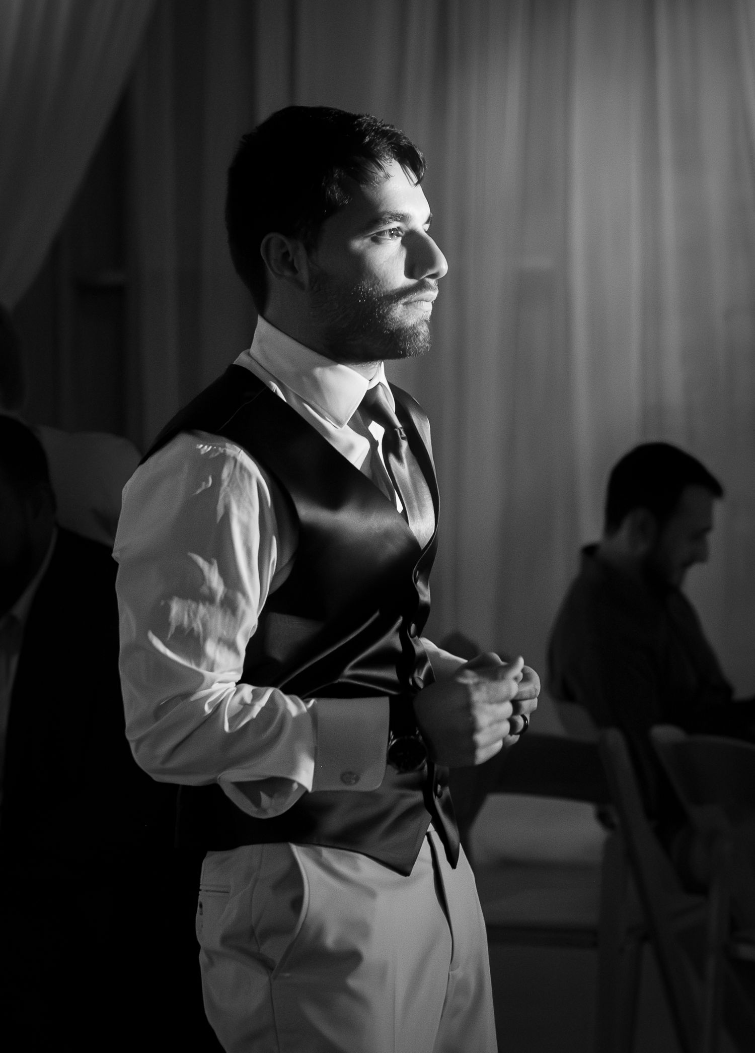 A dapper groom in a black vest and tie stands thoughtfully at a wedding reception in this elegant black and white photo.