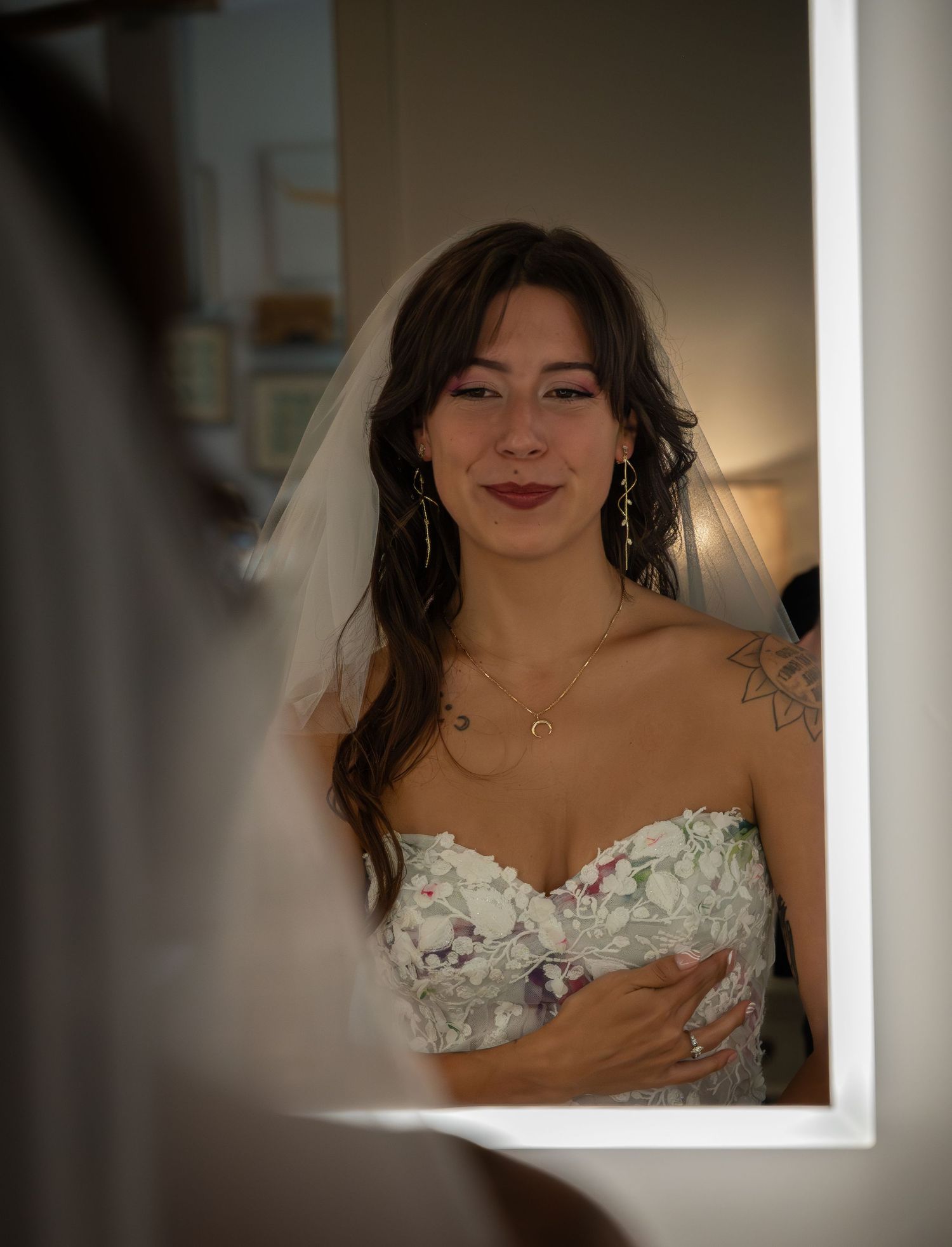 Bride in strapless floral wedding dress and veil smiling at her reflection in a mirror.
