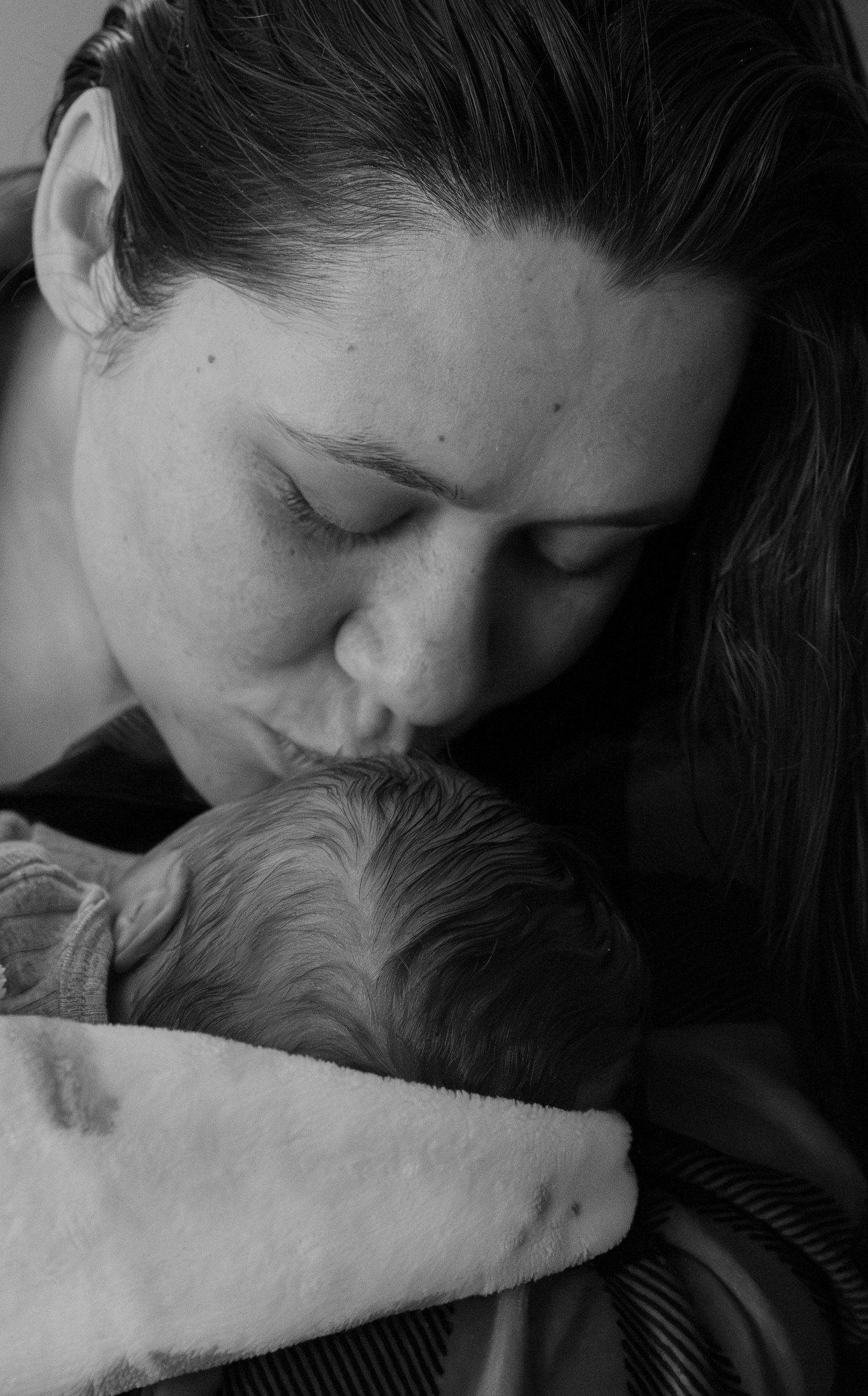 Black and white photo of a mother tenderly kissing her newborn baby's head while cradling it in her arms.