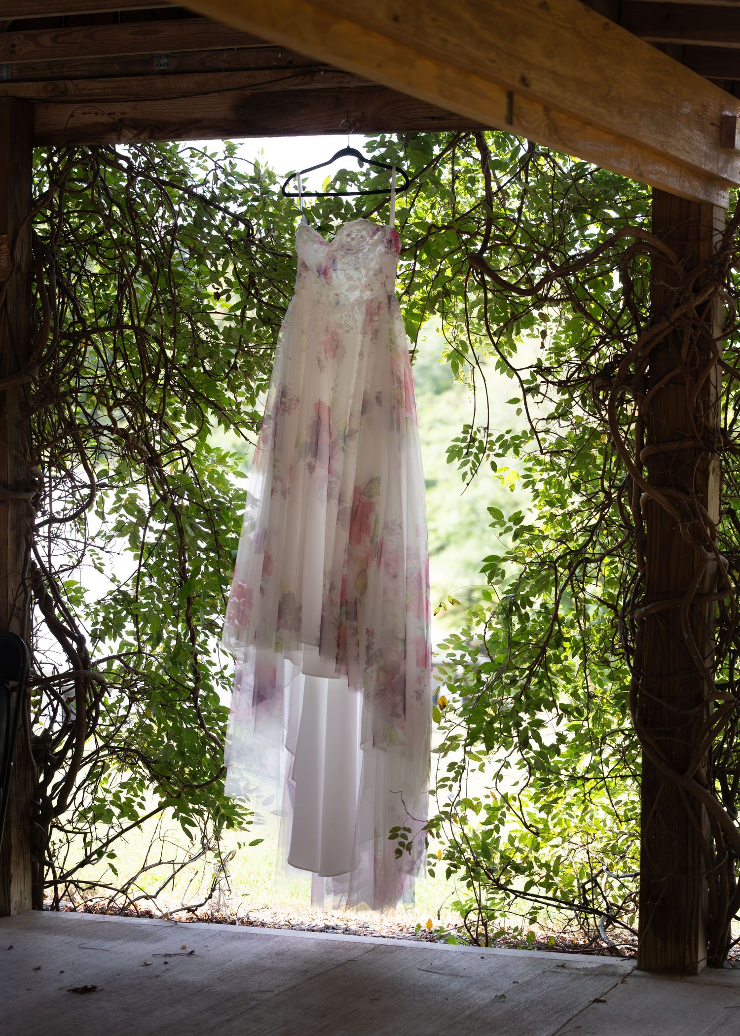 A beautiful floral wedding dress hanging in a rustic vine-covered doorway surrounded by lush green foliage.