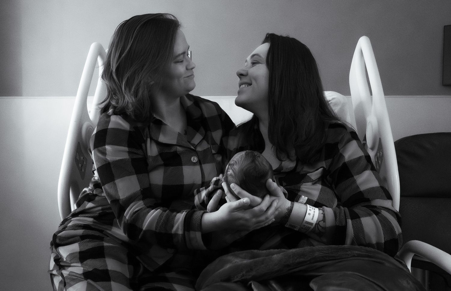 Black and white photo of two women in matching plaid shirts sharing a tender moment with a newborn baby in a hospital room.
