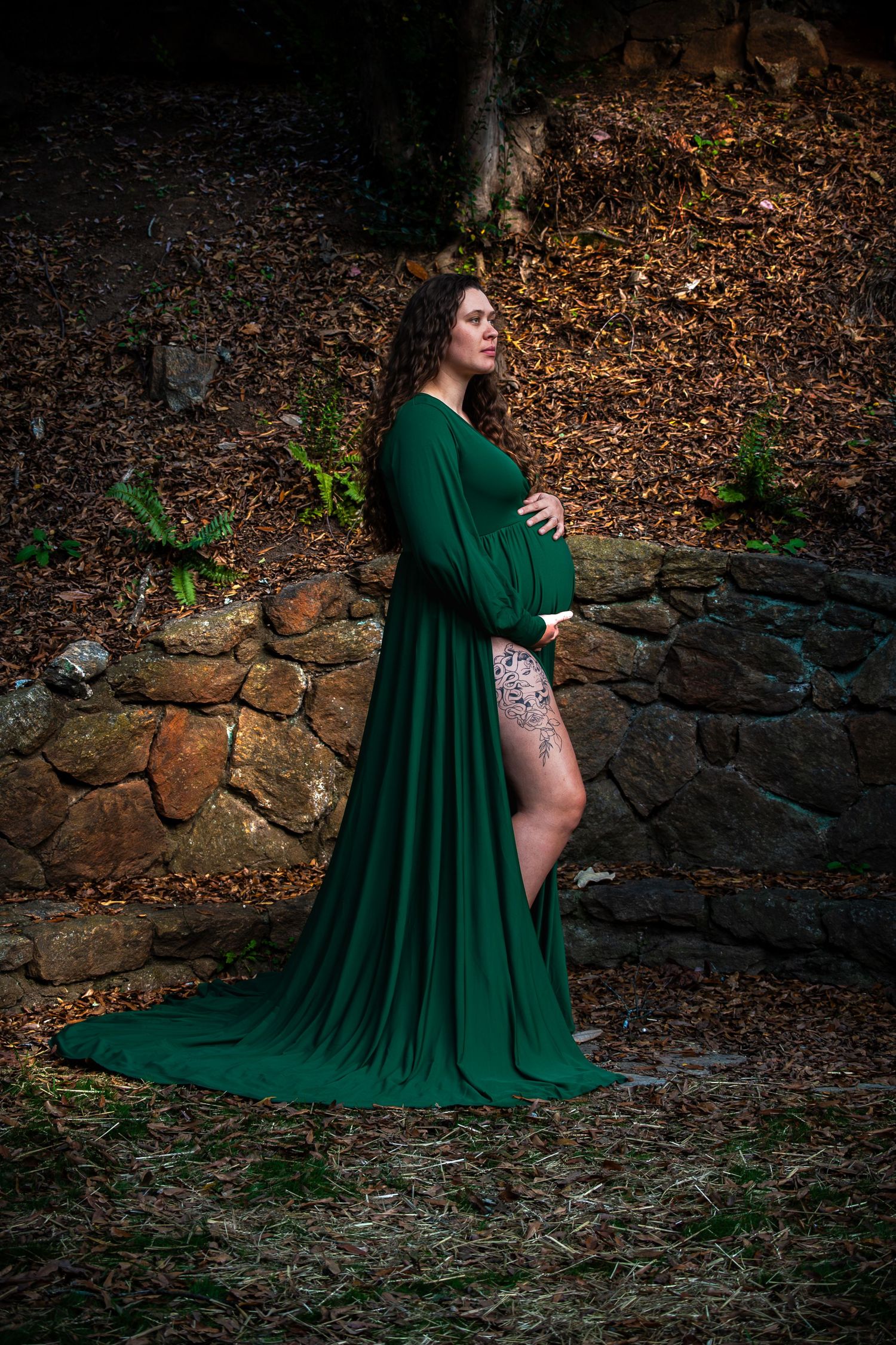 Pregnant woman in elegant green maternity gown poses beside a stone wall in a forest.