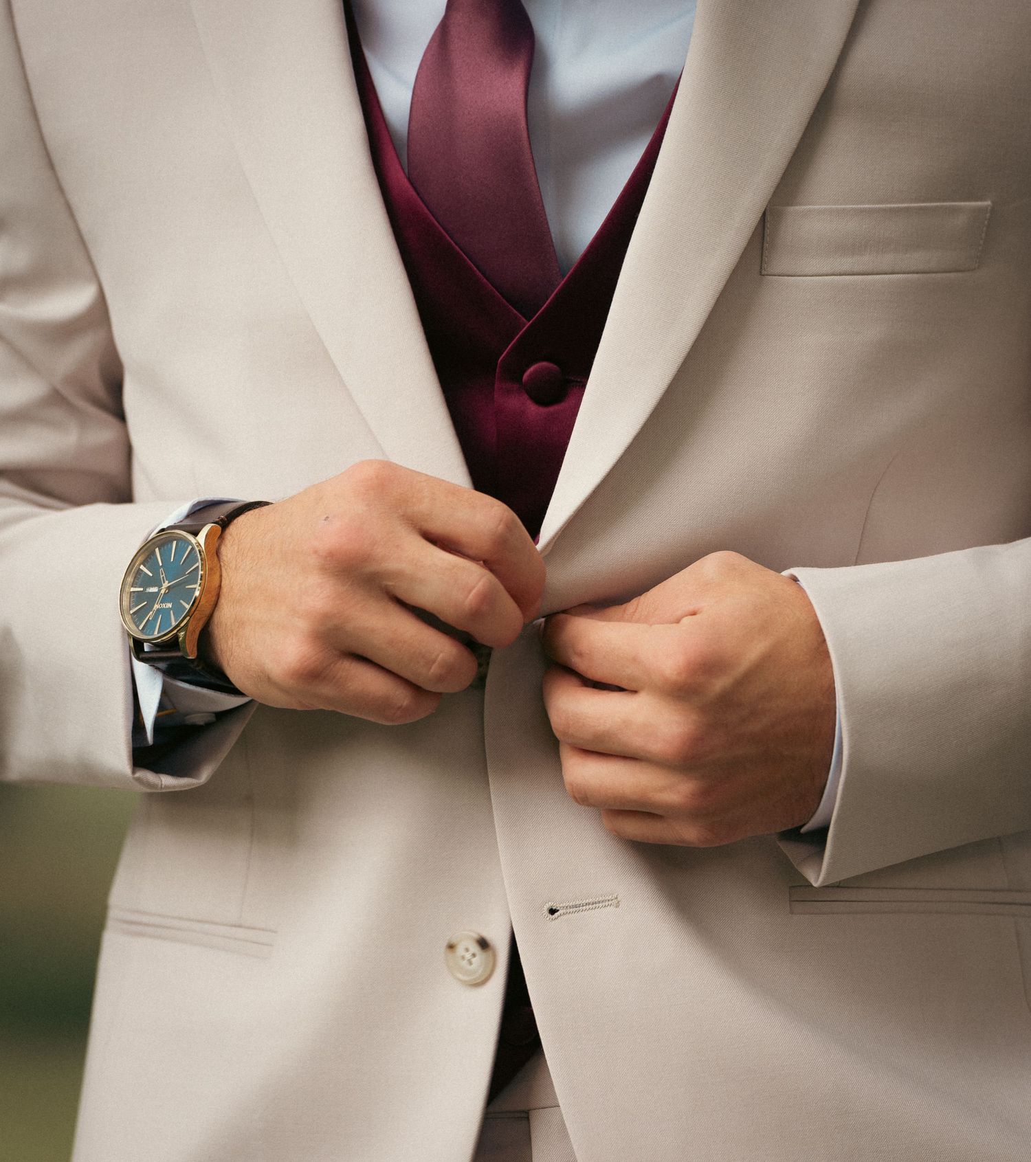 Close-up of groom in beige suit with burgundy vest and tie, adjusting jacket button while wearing a gold watch.
