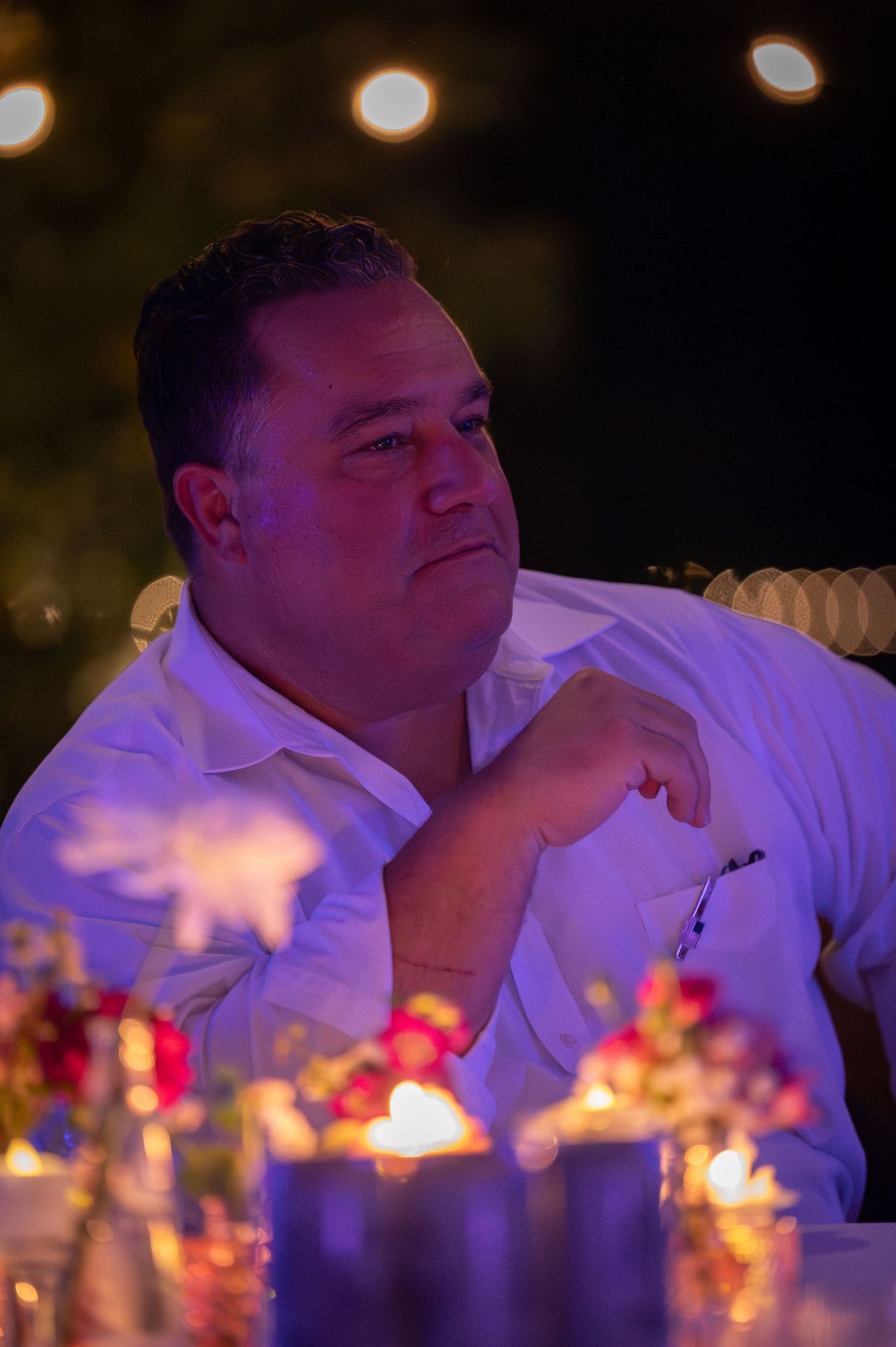 Man in white shirt sitting at candlelit outdoor evening dinner table, gazing thoughtfully into the distance.