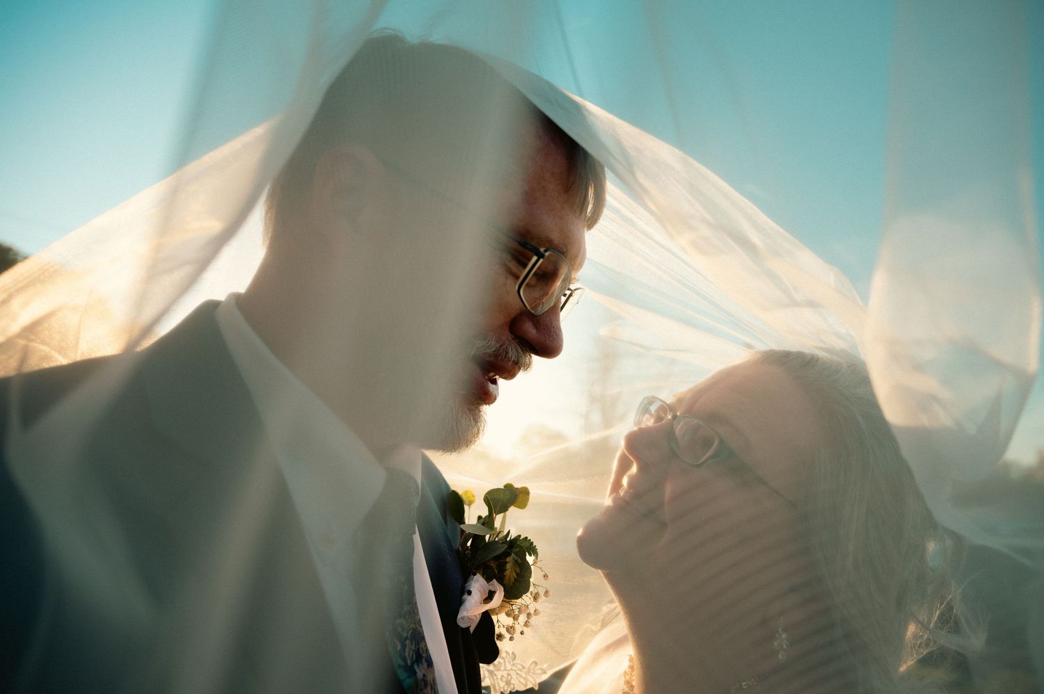 Romantic wedding couple sharing an intimate moment under a flowing white veil with golden backlight.