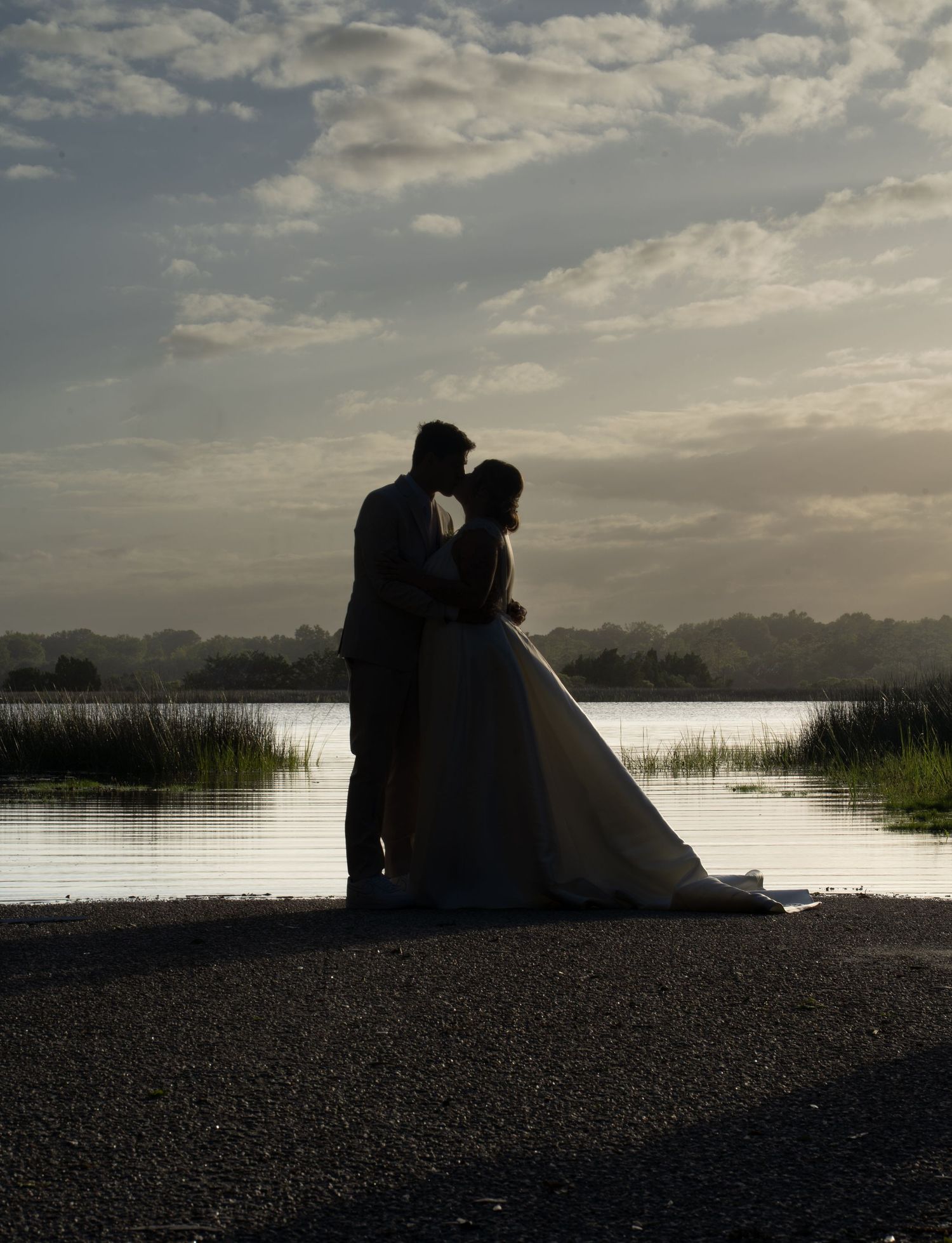 Silhouette of a bride and groom embracing near a tranquil lake at dusk with a cloudy sky backdrop.