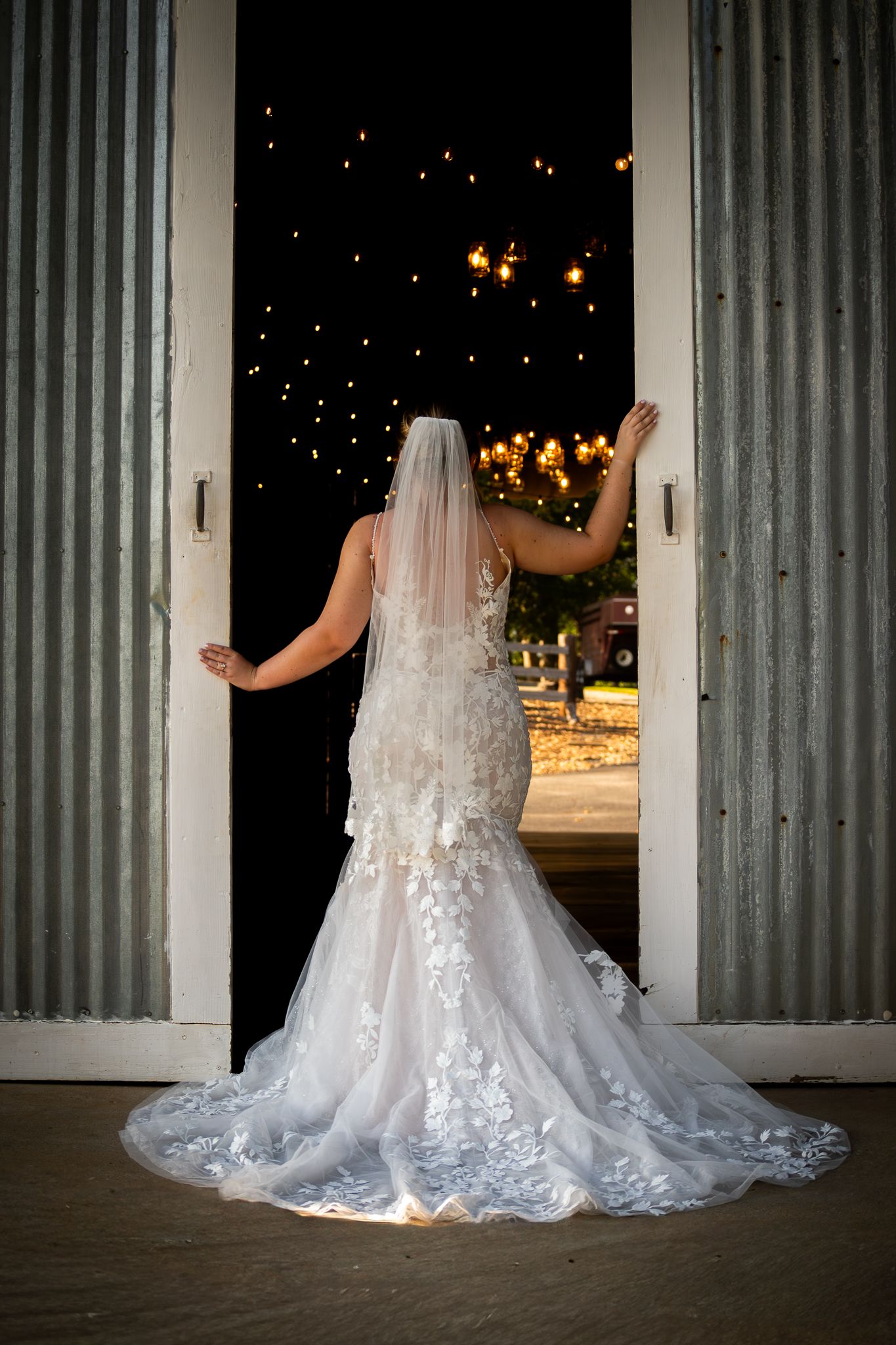 Bride in lace mermaid gown and veil stands in barn doorway, gazing at twinkling string lights outside.