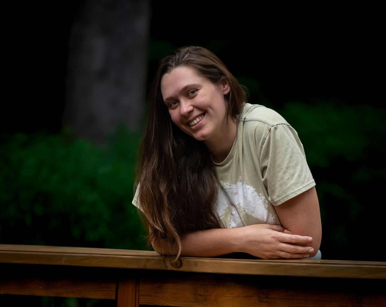 Young woman with long brown hair smiling warmly while leaning on a wooden railing outdoors.