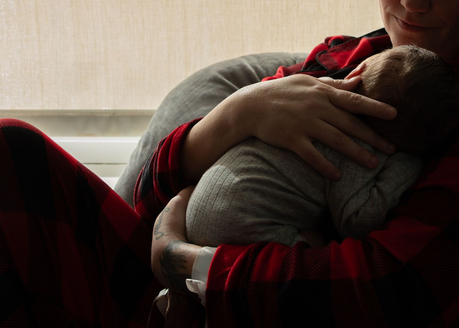 Adult in red plaid shirt tenderly cradling a sleeping newborn baby in gray onesie in a dimly lit room.