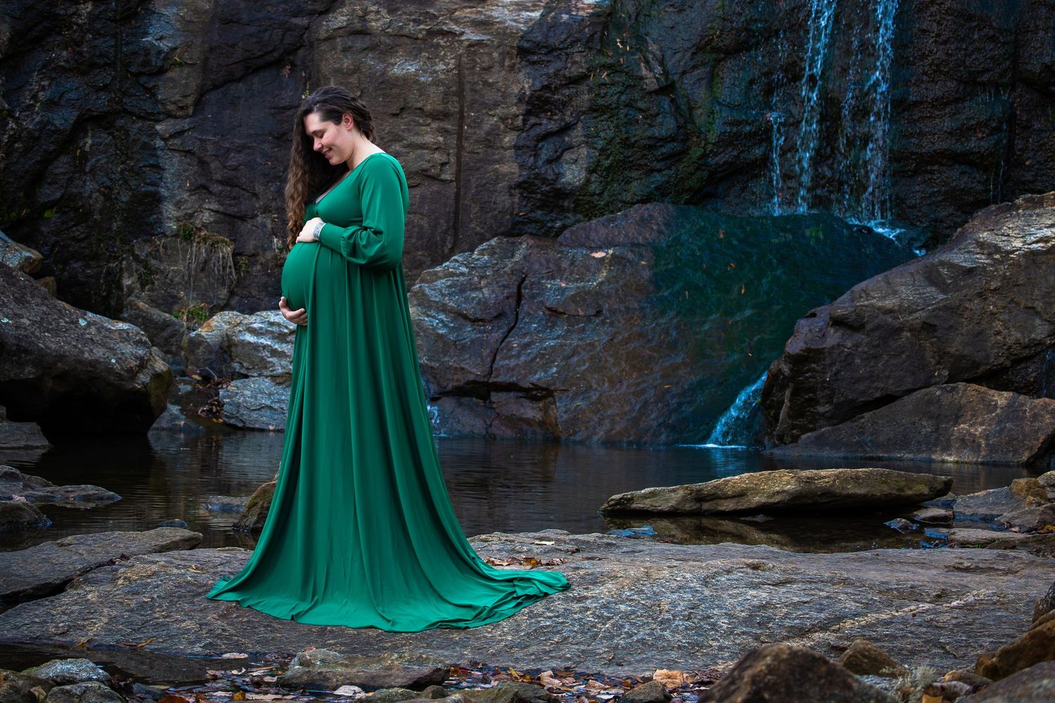 Pregnant woman in elegant green gown poses on rocky terrain near a waterfall during a maternity photo shoot.