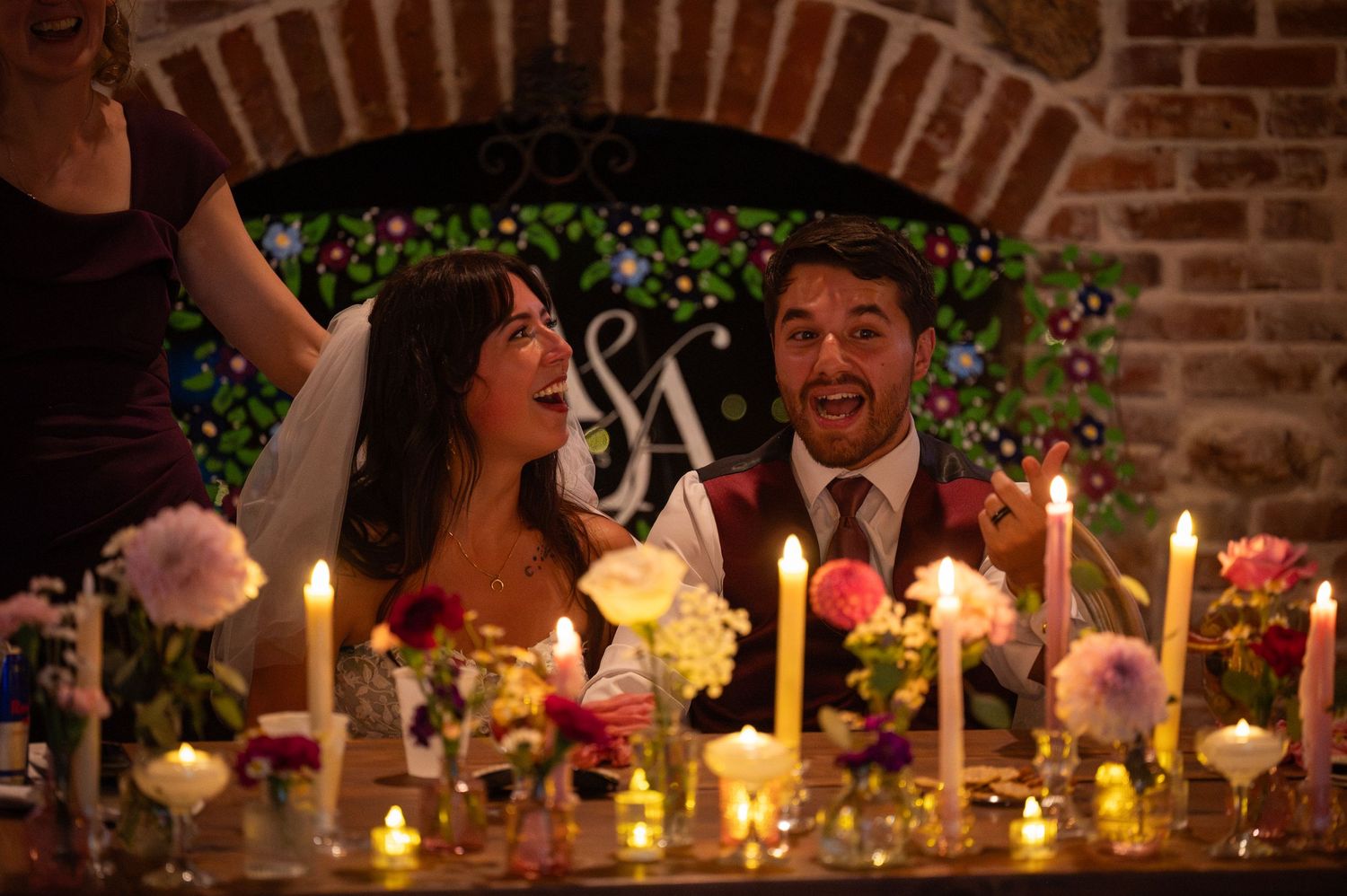 Bride and groom laughing joyfully at candlelit wedding reception table with floral arrangements and monogram backdrop.
