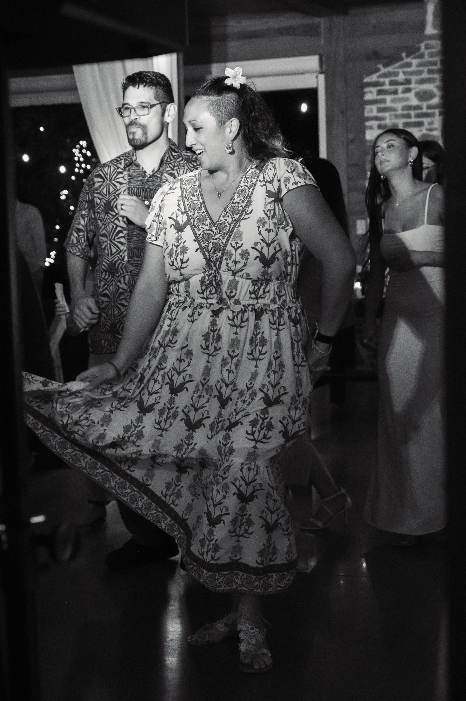 Black and white photo of a woman in a floral dress dancing joyfully at an indoor event with guests in the background.