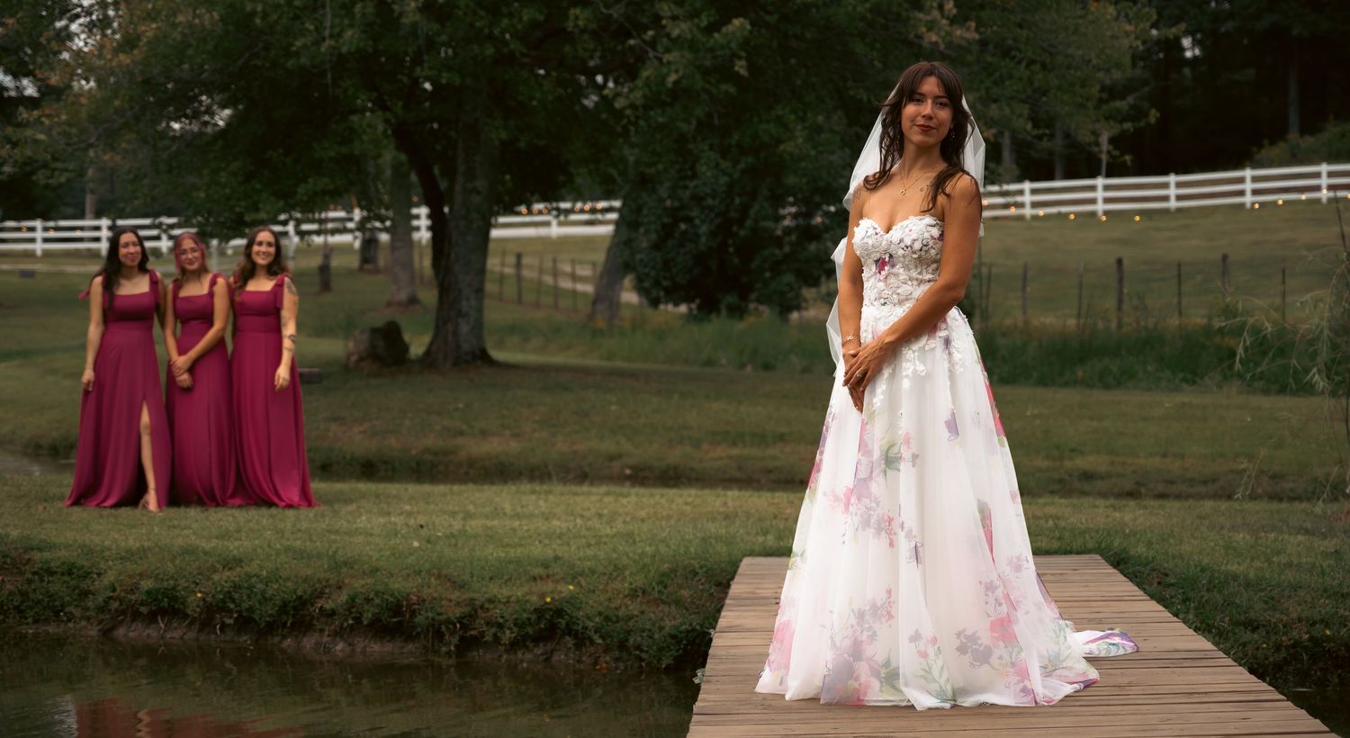 Stunning bride in floral wedding gown poses on dock while bridesmaids in burgundy dresses stand behind.