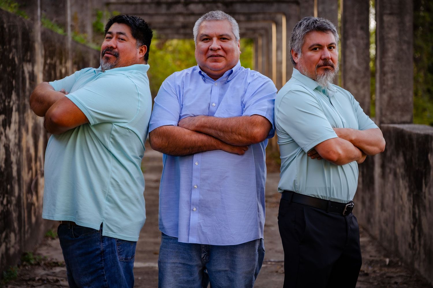 Three men in light blue shirts posing with arms crossed outdoors.