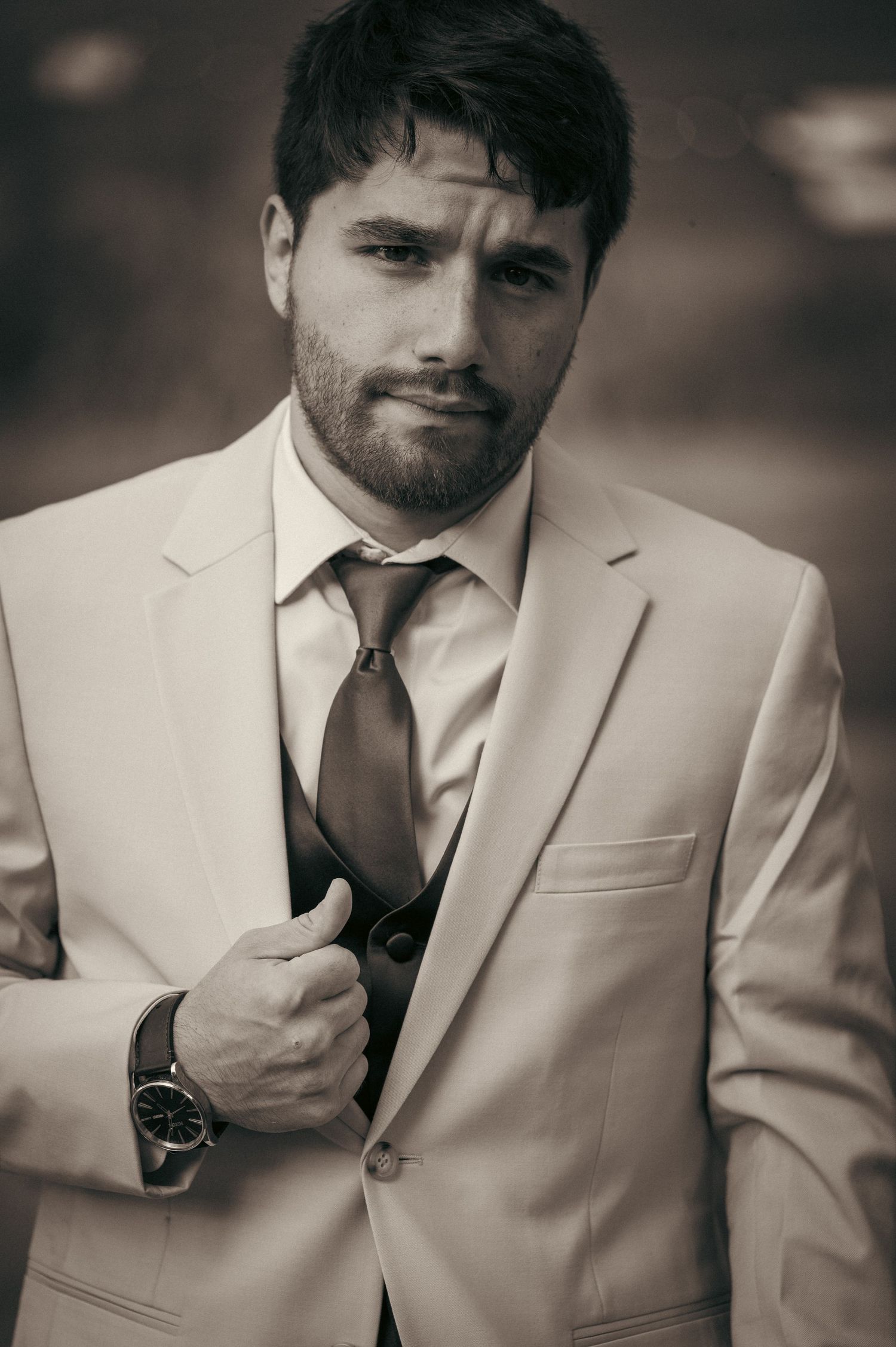 Sepia-toned portrait of bearded groom in white suit and tie, posing confidently outdoors.