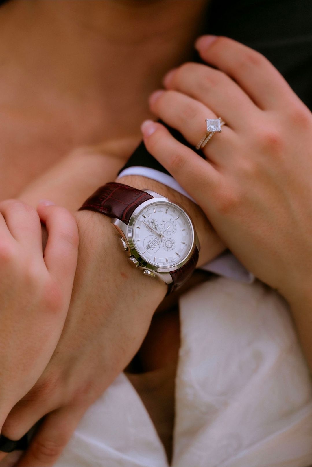 Close-up of a bride's hand with a diamond engagement ring resting on a groom's wrist wearing a brown leather watch.