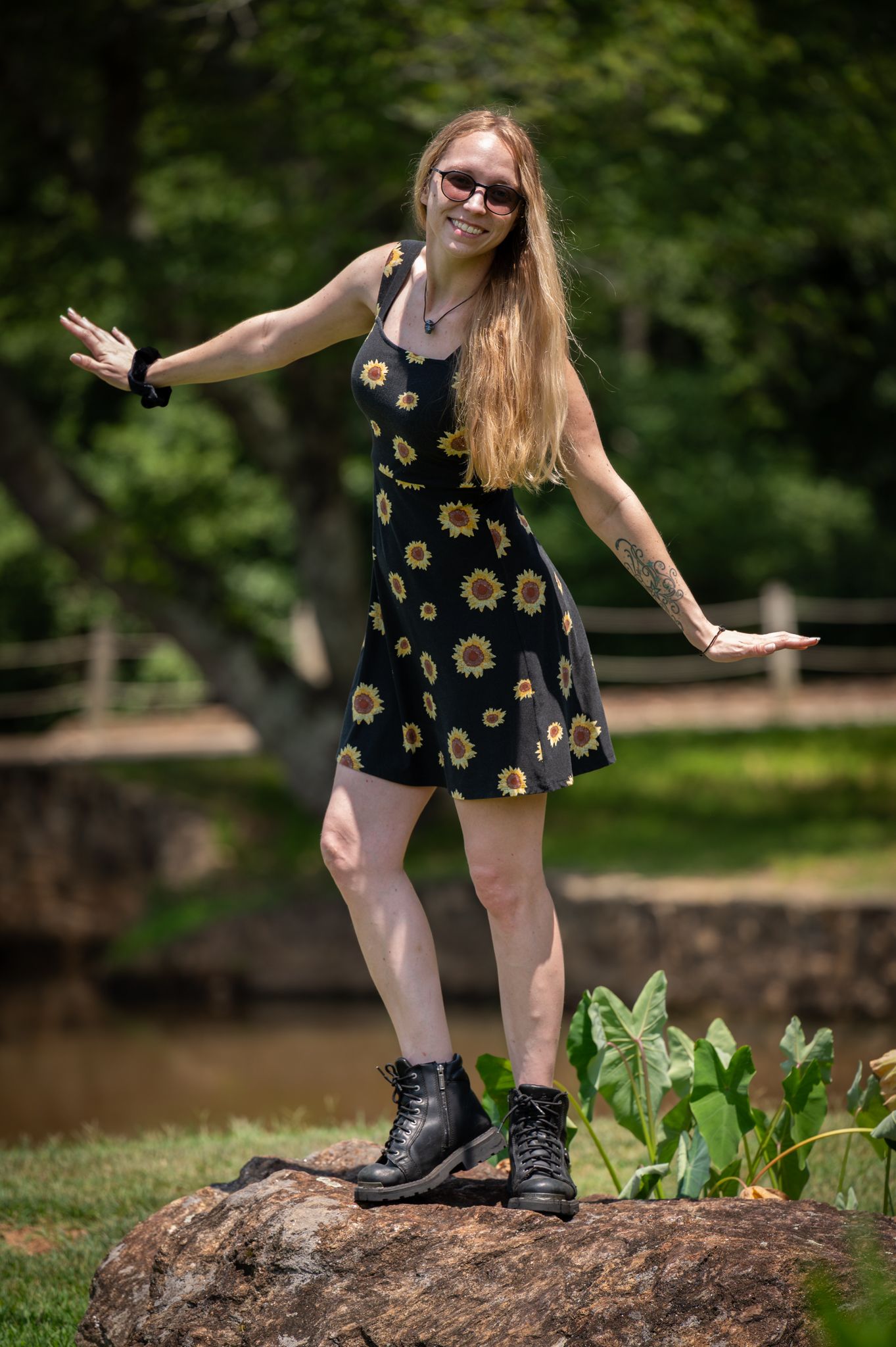 Young woman in sunflower dress and combat boots balancing on a rock in a lush green park setting.
