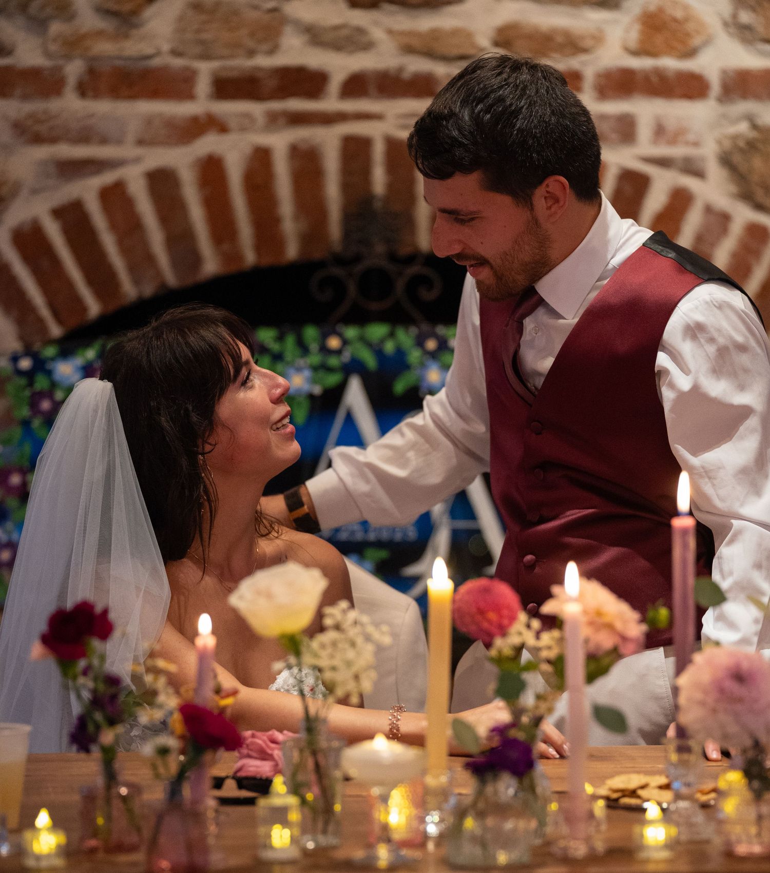 Bride and groom share a romantic moment at candlelit wedding table adorned with colorful flowers in a rustic stone venue.